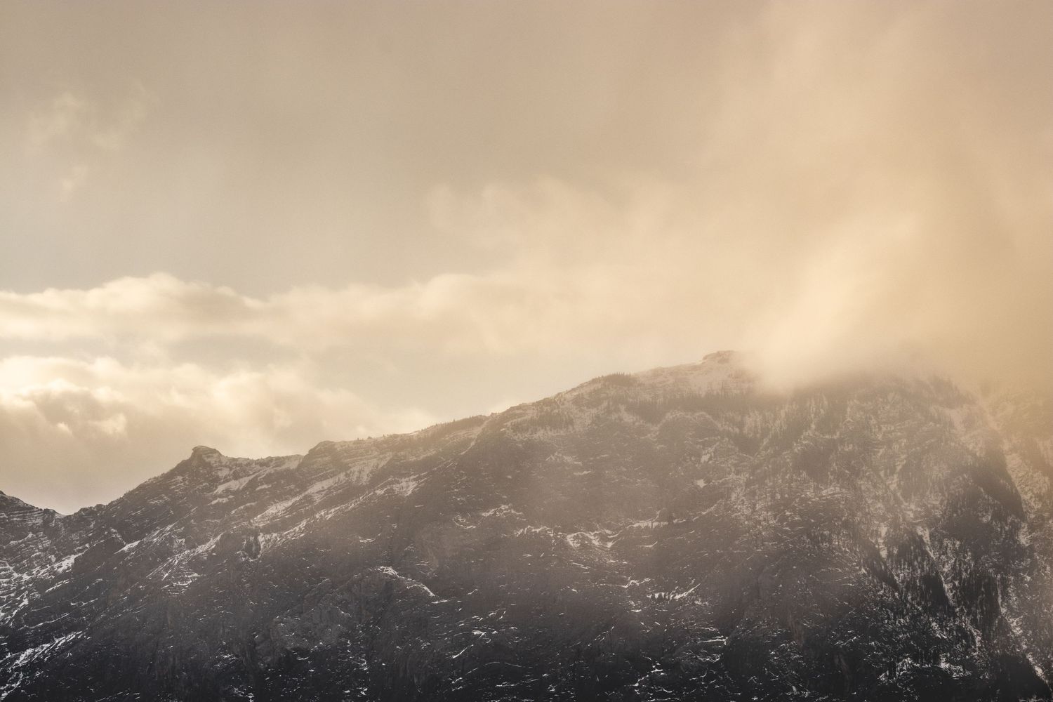 Dramatic cloudy sunset illuminates snow-capped mountain peaks in muted golden tones.