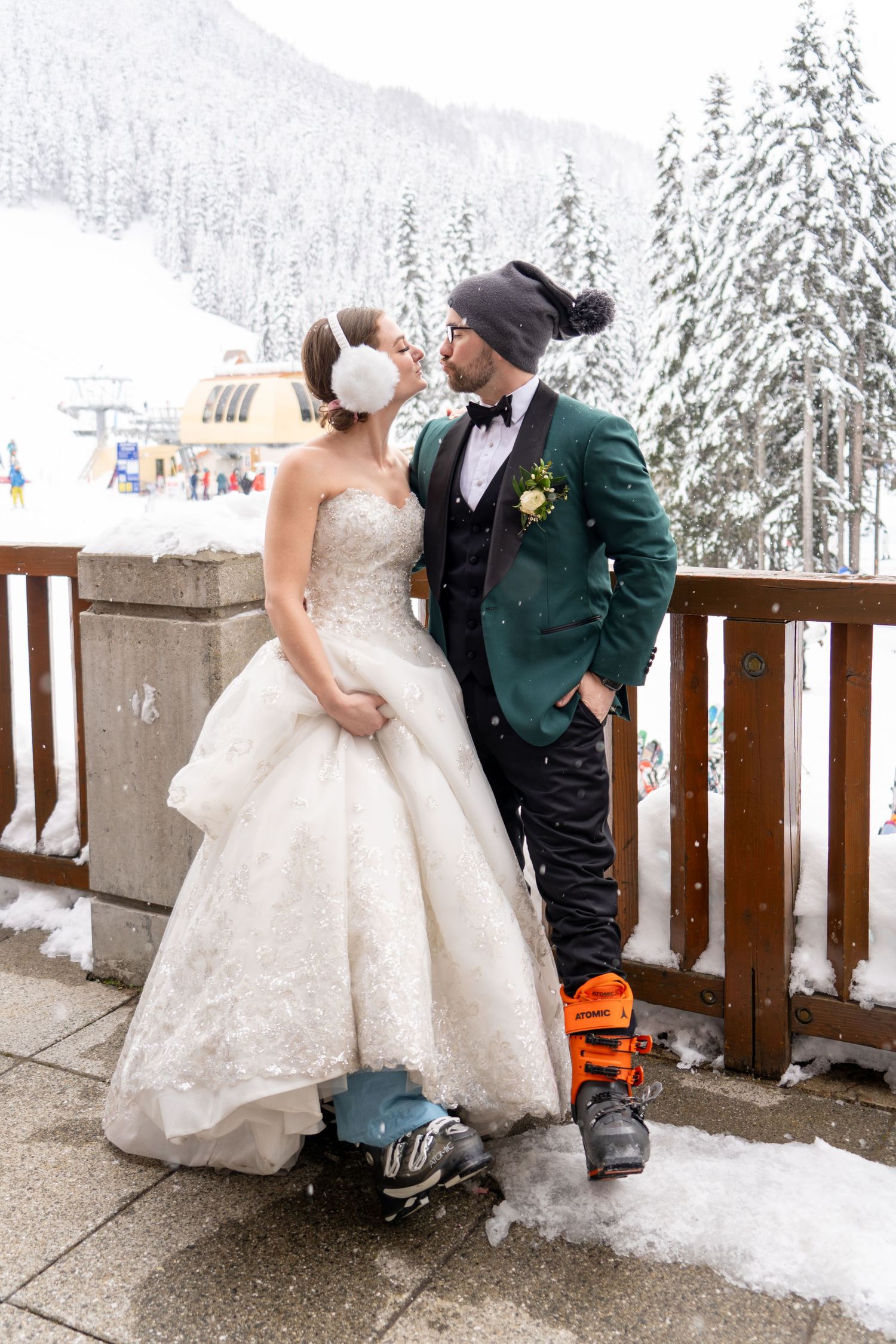 A winter wedding couple in ski gear poses on a snowy deck in front of evergreen trees.