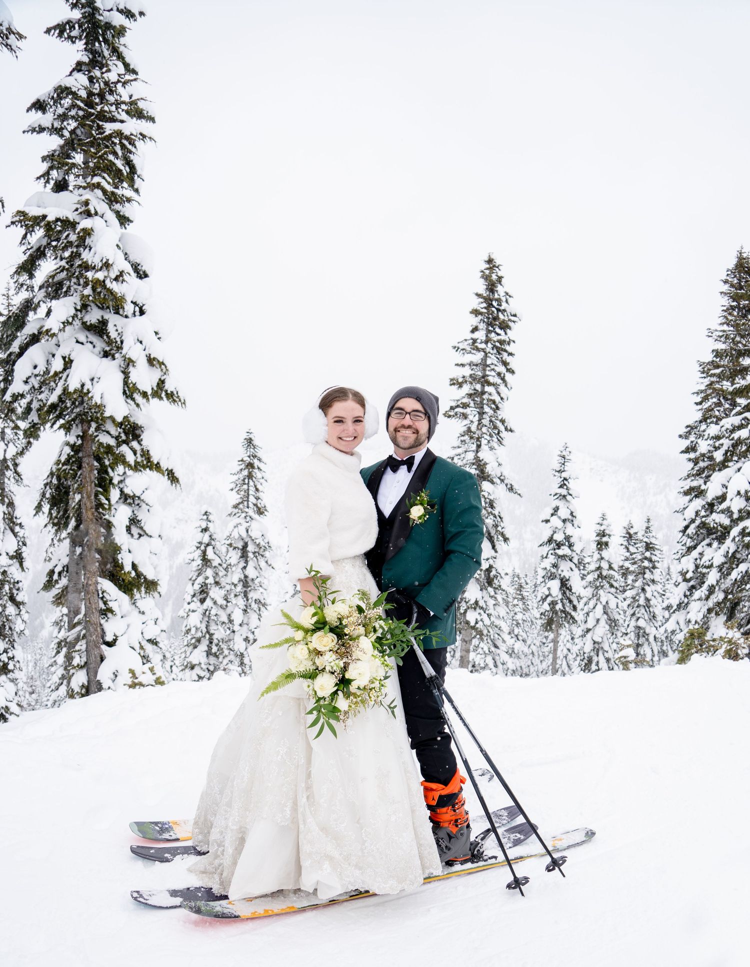 A newlywed couple poses on skis in front of snow-covered pine trees at a winter resort.
