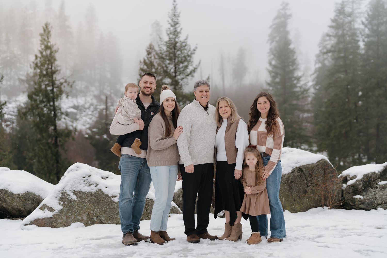 Family of seven posing together in a snowy winter landscape with pine trees and fog in the background.