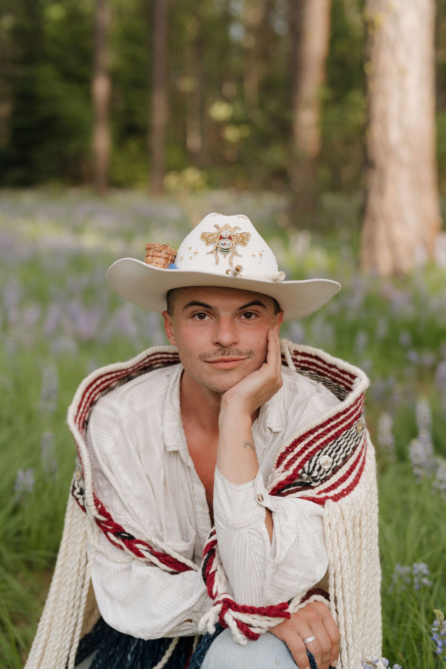 Man in white cowboy hat and shirt sitting in rope chair among wildflowers in forest setting.
