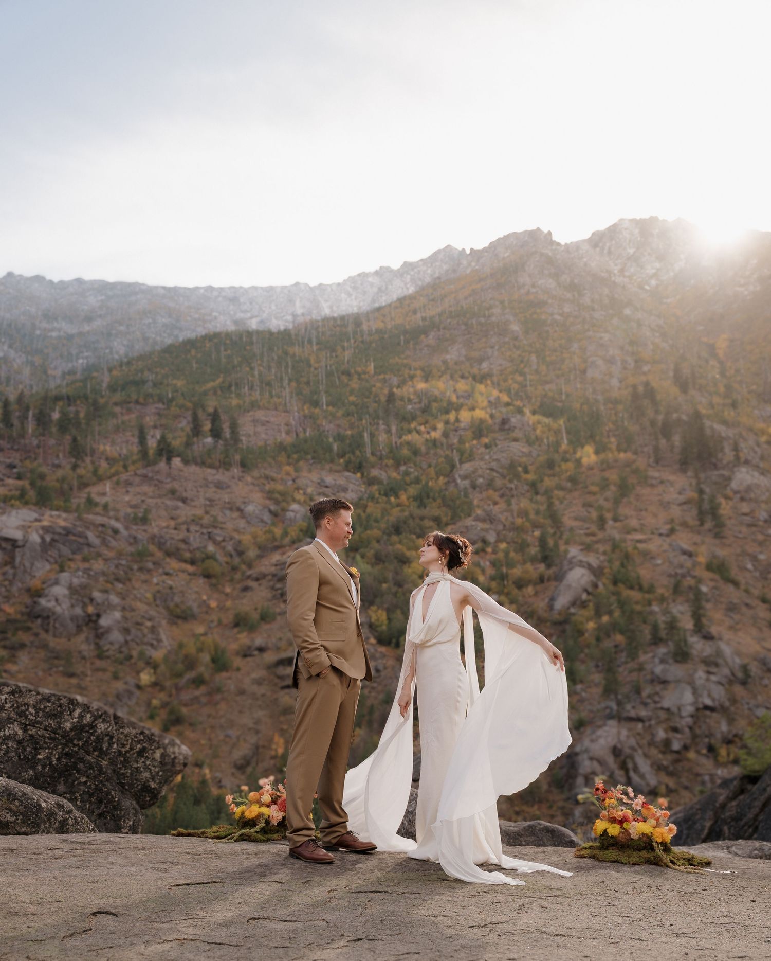 A couple stands together on a mountain ridge overlooking a scenic valley at sunset during their wedding photo session.