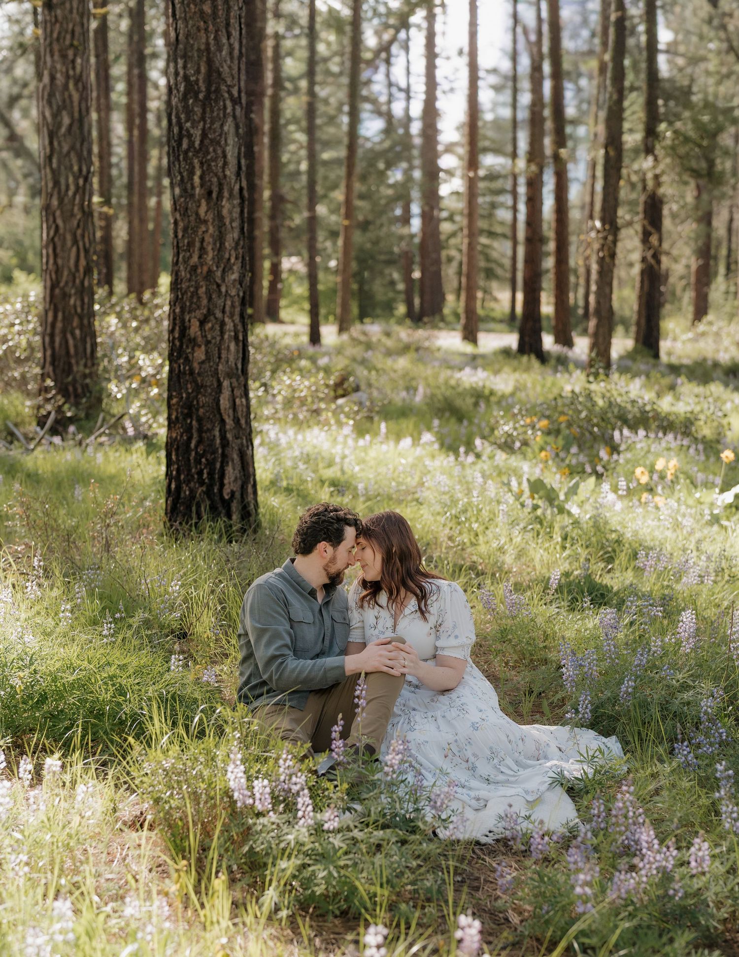 Couple sharing a tender kiss while sitting among wildflowers in a sunlit forest during an engagement session.