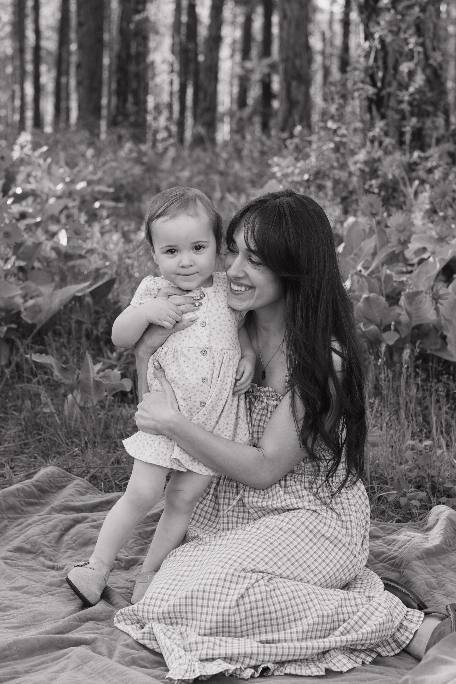Black and white photo of a smiling mother holding her baby girl while sitting on a blanket in a forest.