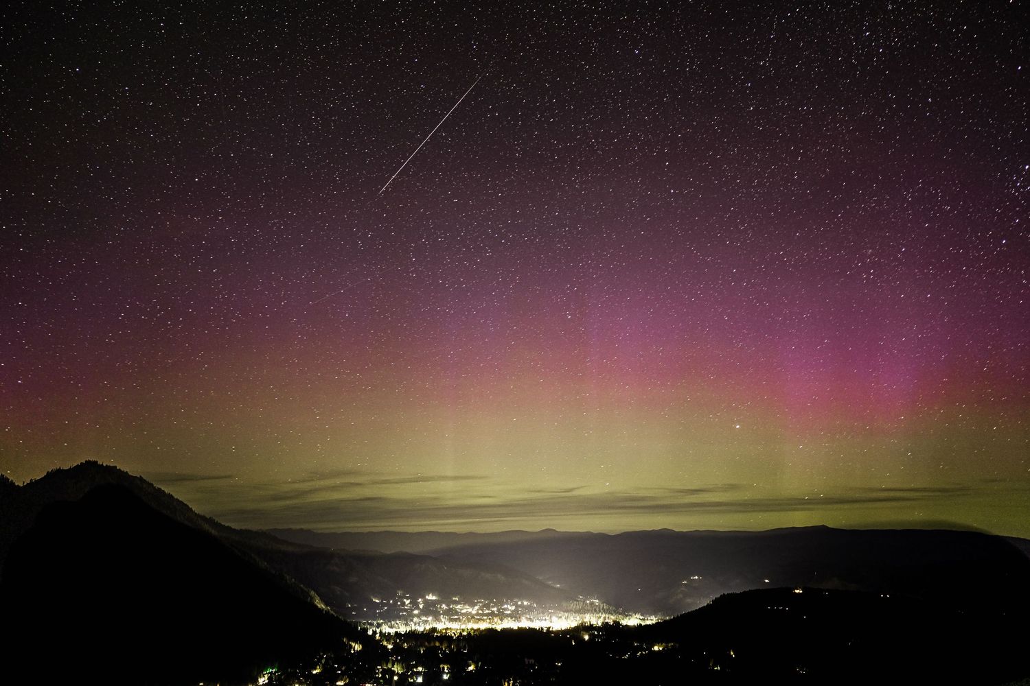 Purple and green aurora borealis illuminates night sky over city lights in valley below.