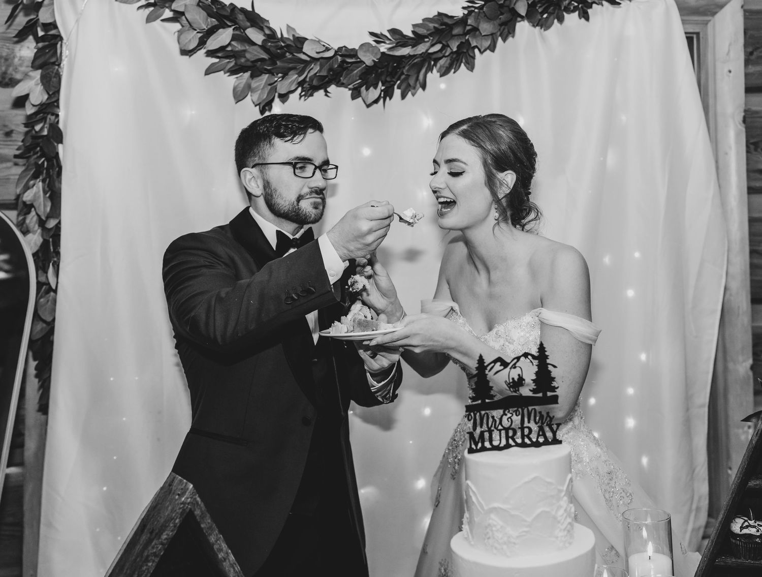 Newlyweds share cake during their wedding reception in front of a decorated floral backdrop.