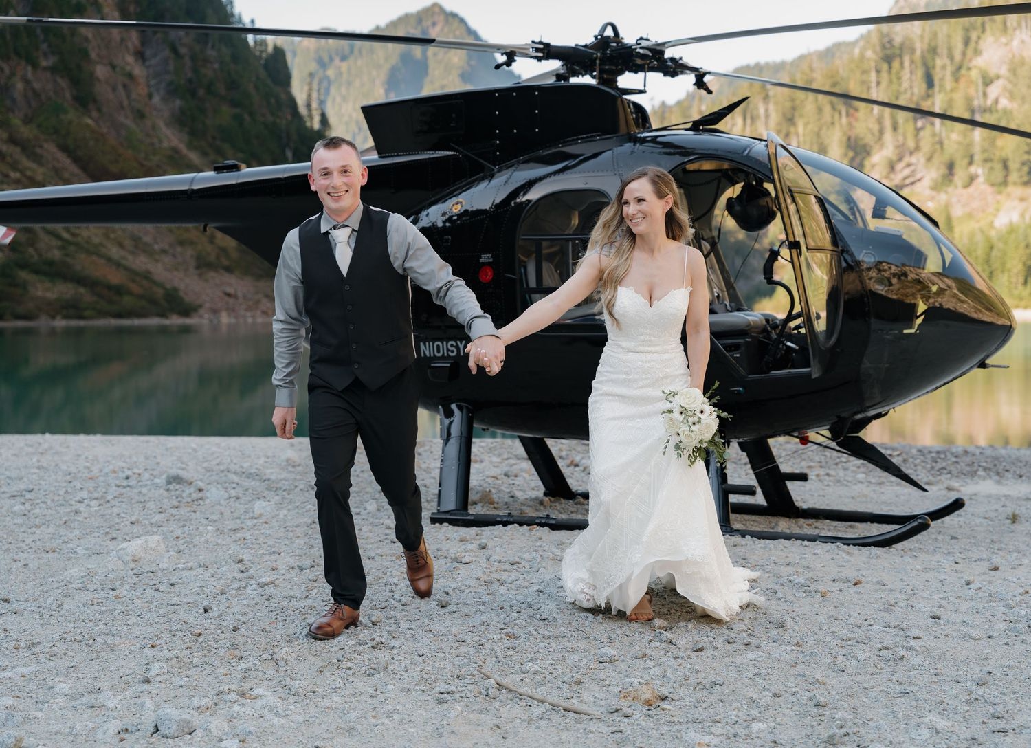 Bride and groom holding hands near black helicopter in scenic mountain setting.