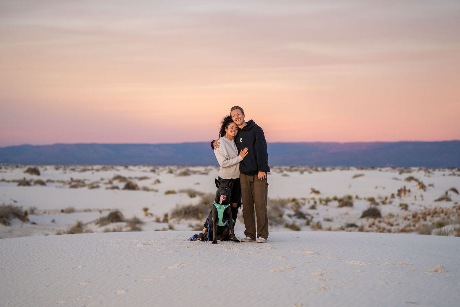 A couple stands together at White Sands National Park during sunset with pink and purple sky in the background.