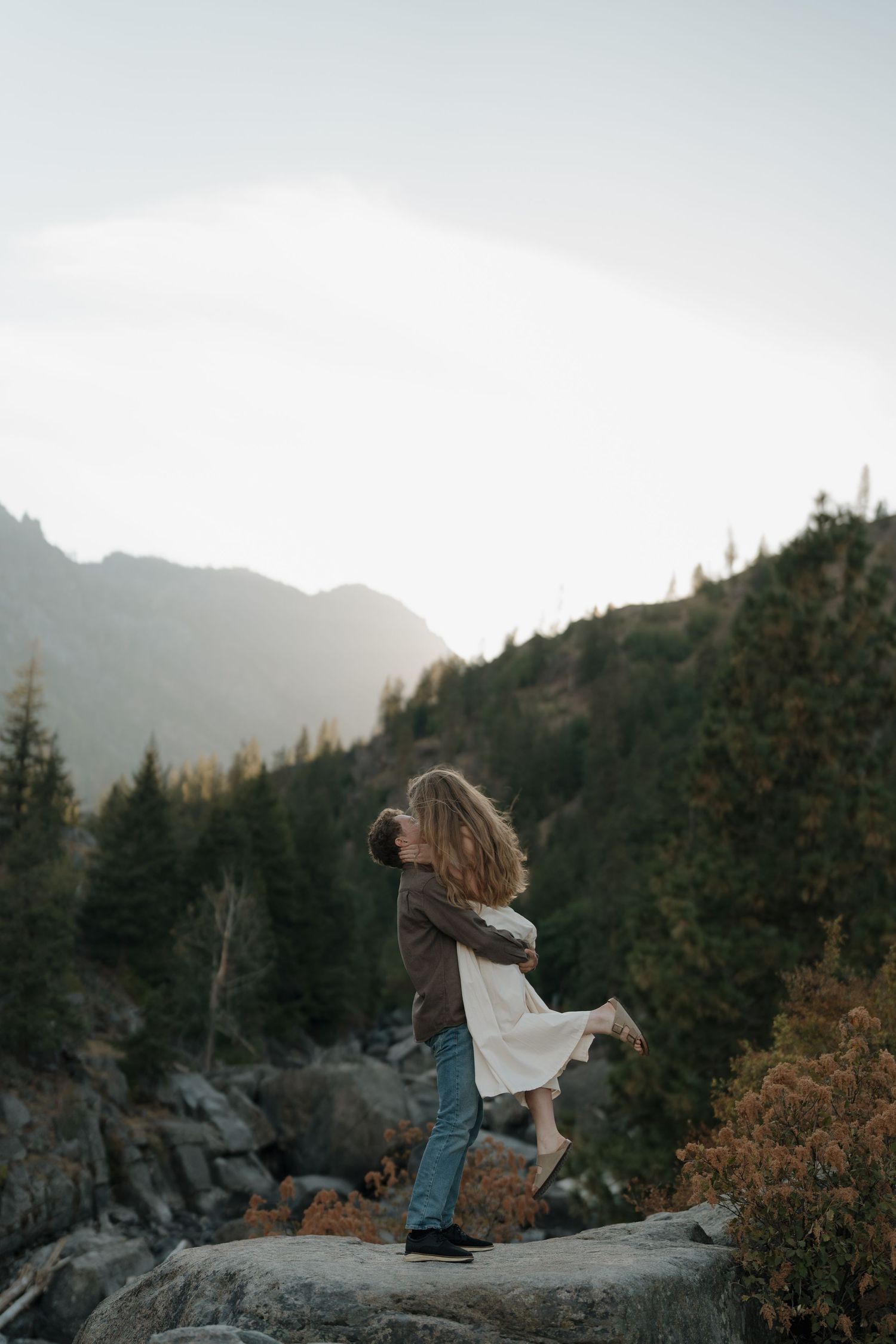 Couple embracing on rocky mountain overlook surrounded by pine trees and misty peaks at golden hour.