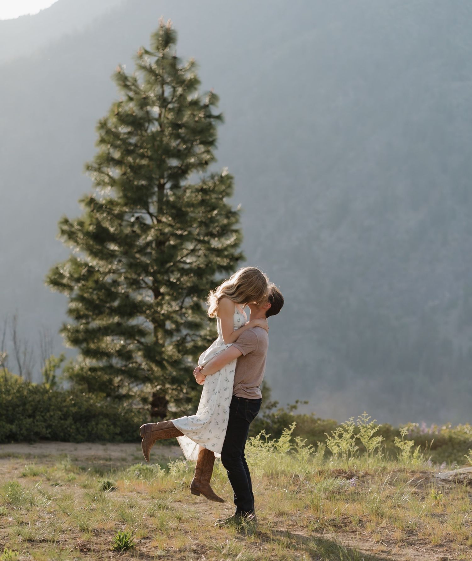 A couple embraces in a romantic moment on a mountainside with a tall evergreen tree and misty peaks in the background.