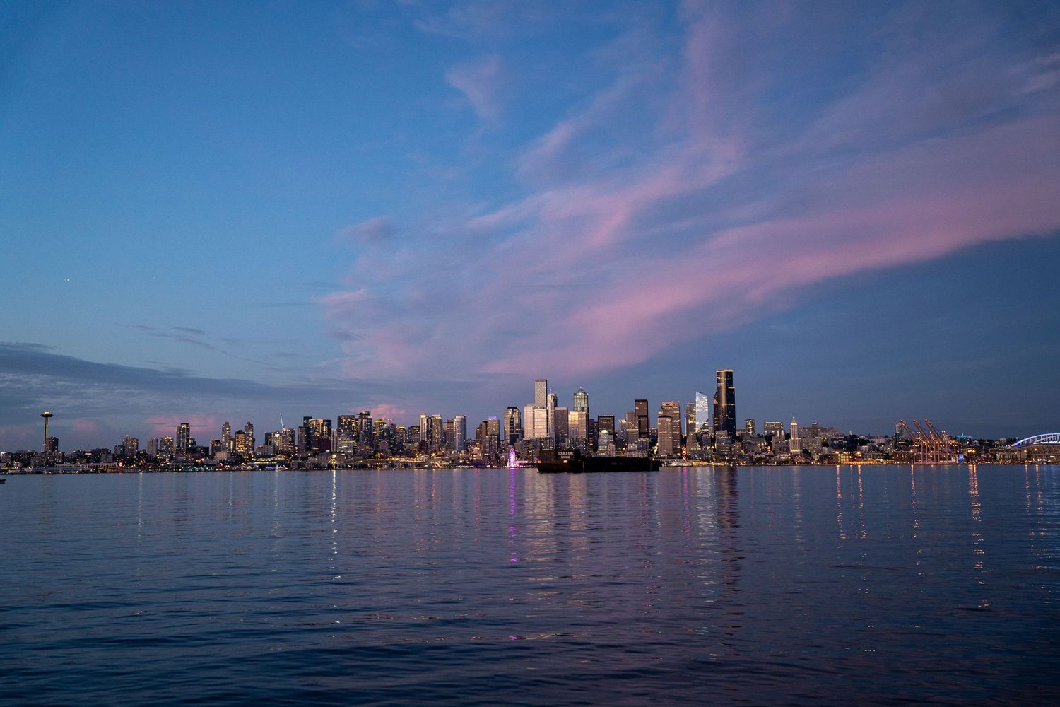 Seattle skyline reflecting on Elliott Bay waters during sunset with pink clouds and blue sky.