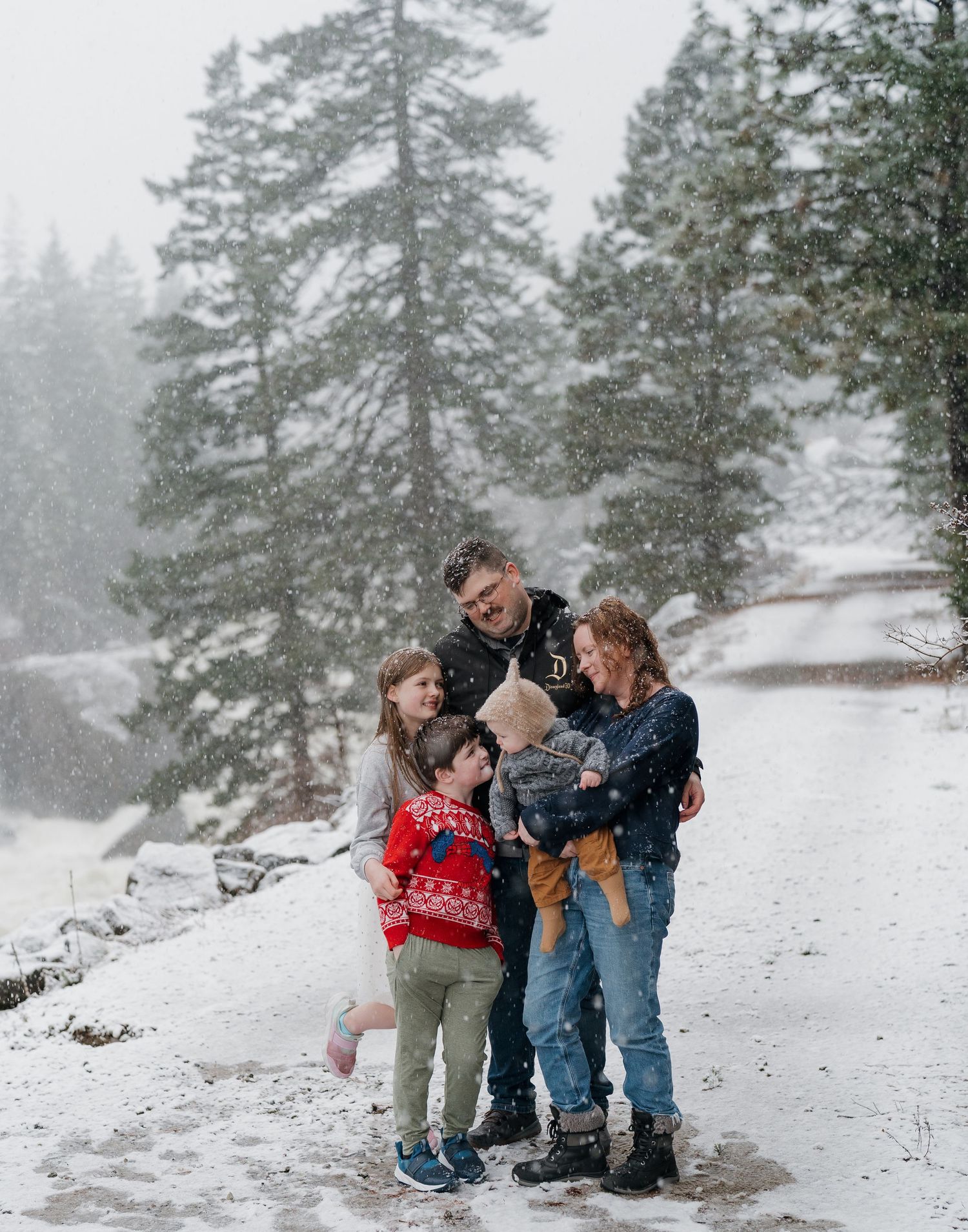 A family of five hugs and laughs together in a snowy evergreen forest while snow falls, with children in cozy winter sweaters.