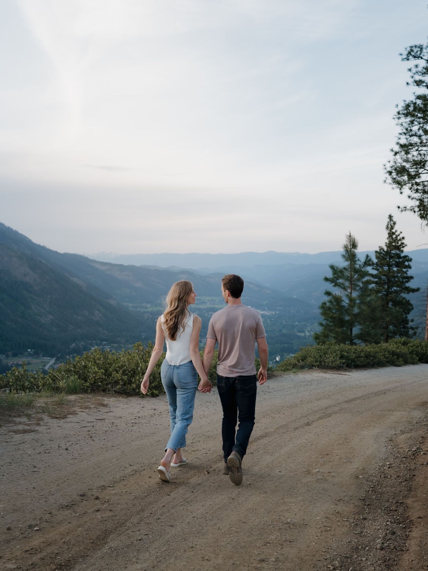 A couple walks along a mountain trail with sweeping valley views at sunset, holding hands and enjoying the scenic vista.