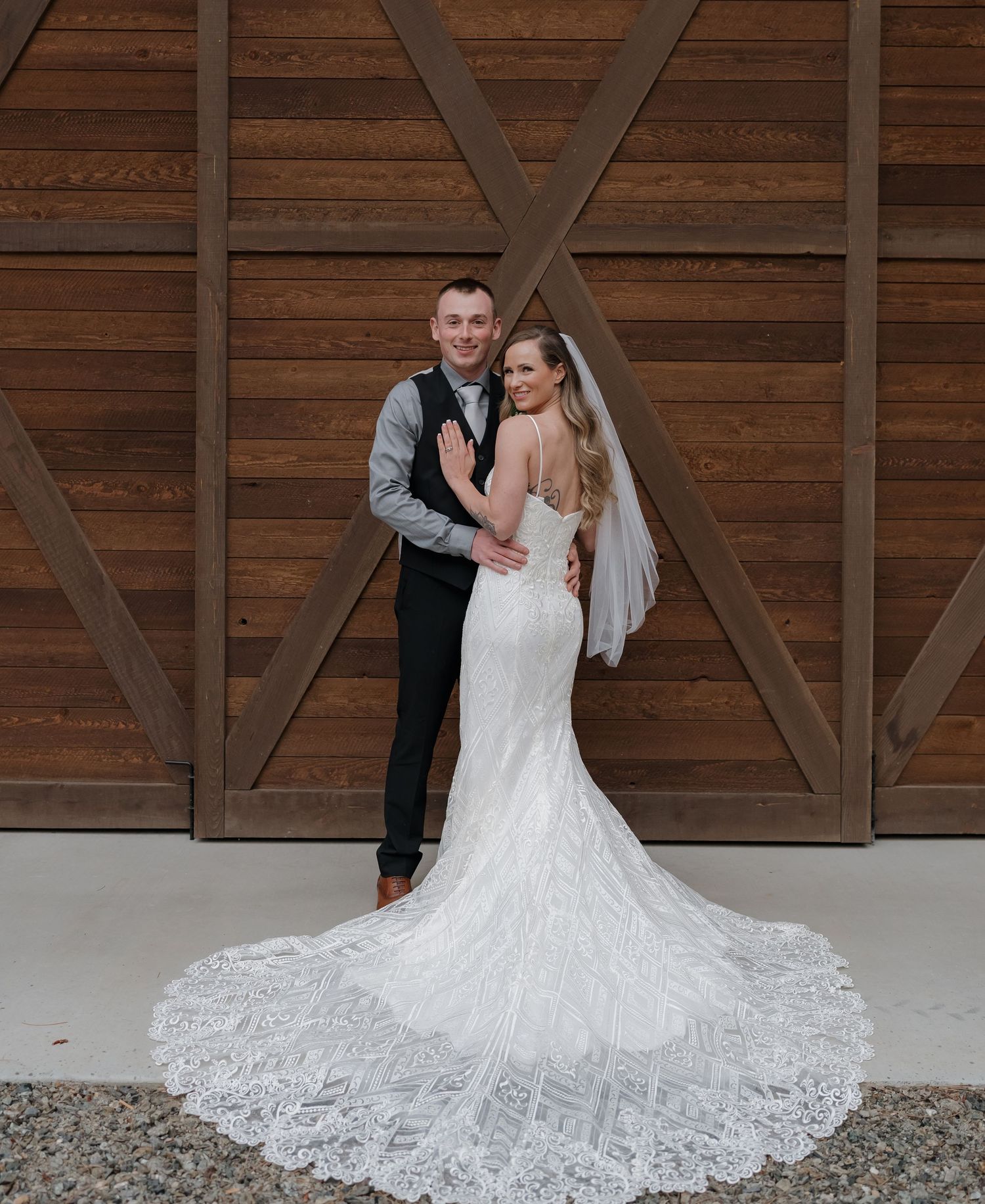 Newlyweds posing against rustic wooden barn doors with bride in lace train wedding dress.