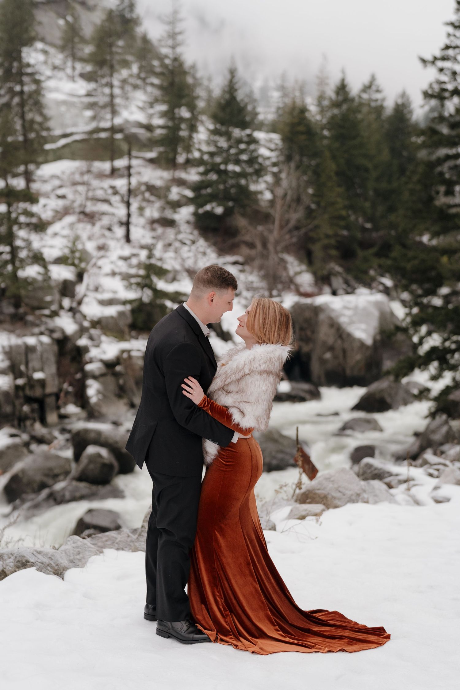 Couple embracing in snowy winter landscape, woman in rust velvet gown with fur wrap, man in black suit, evergreen trees behind.