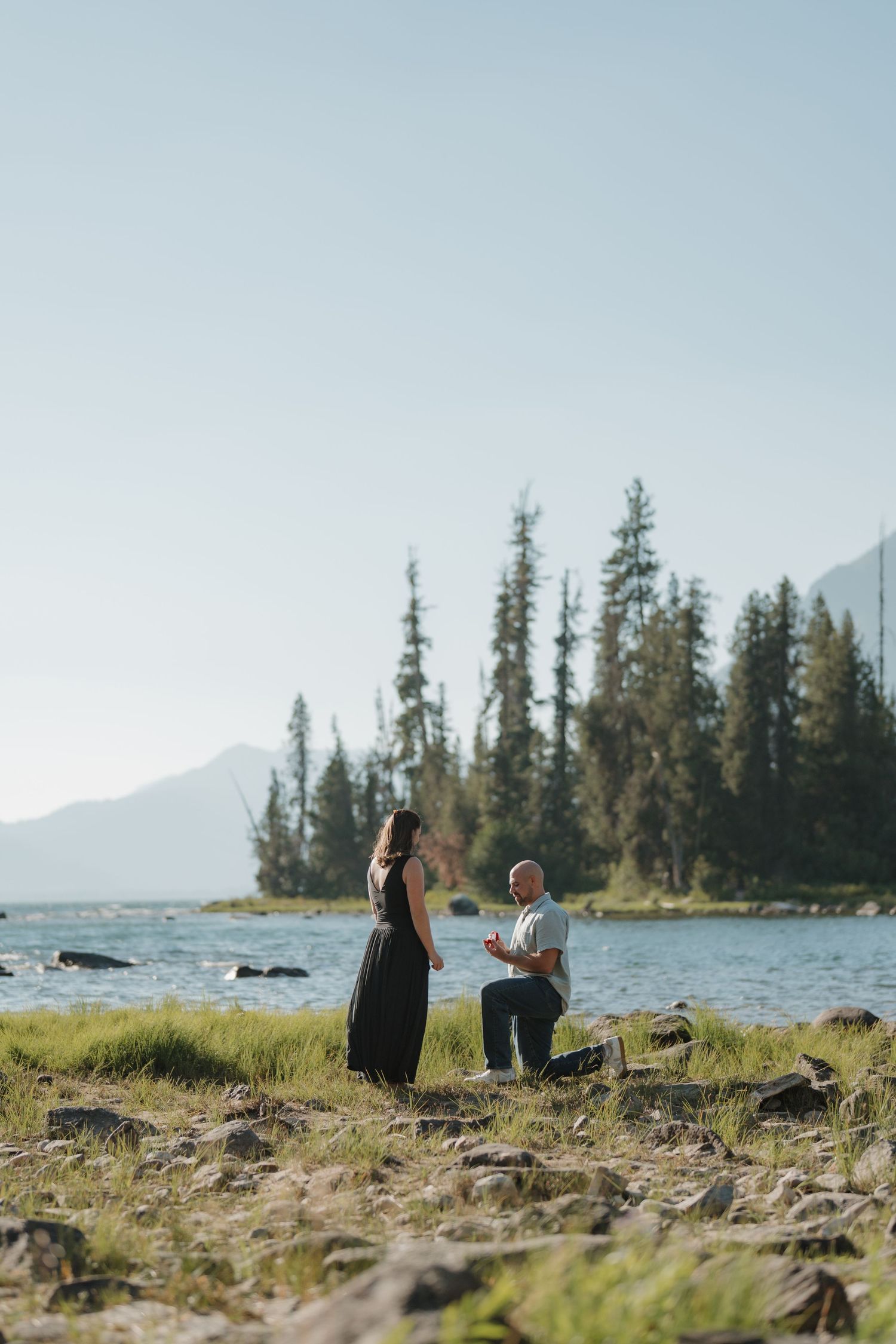 Man proposes on one knee to woman by a scenic mountain lake surrounded by pine trees on a sunny day.