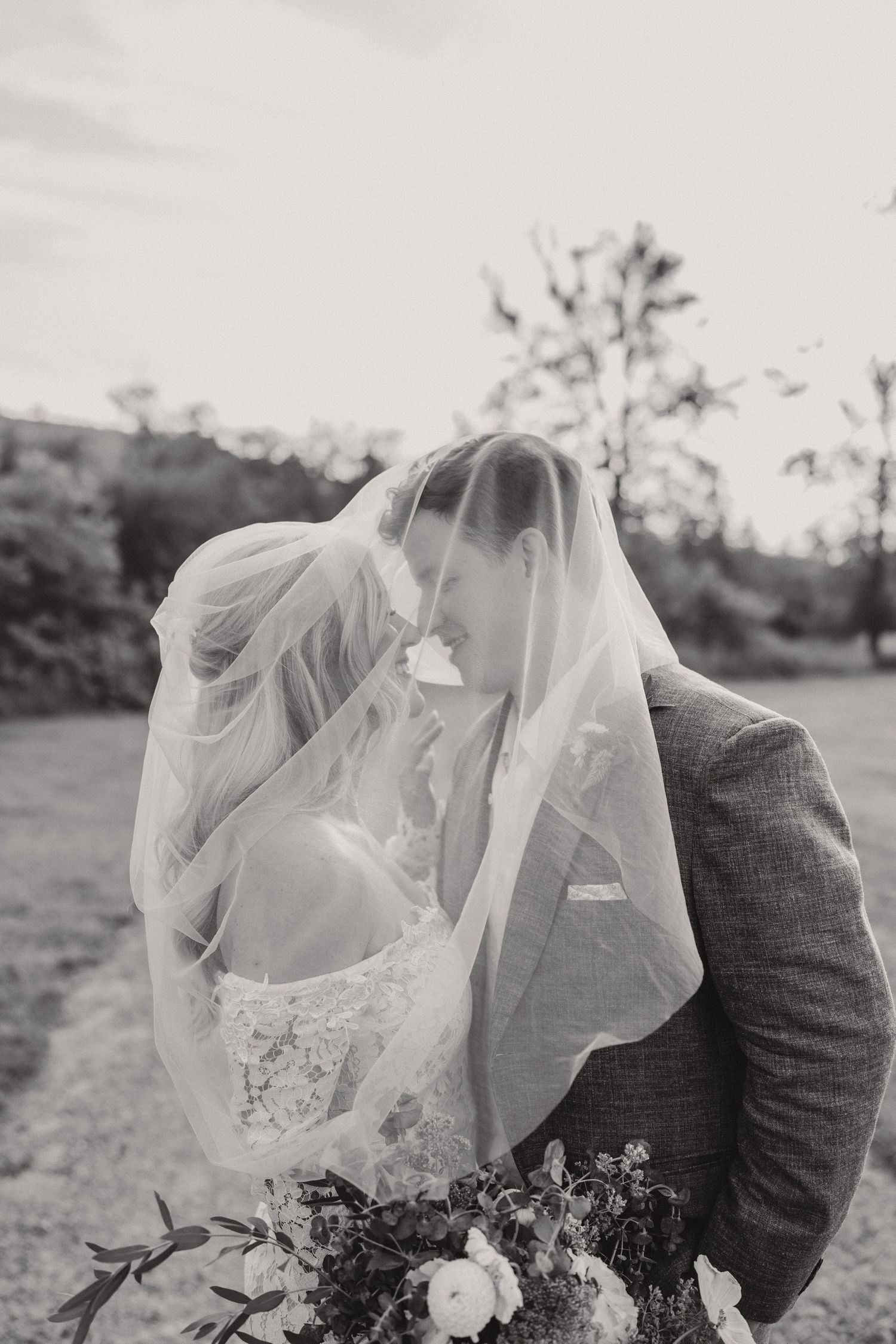 A romantic black and white wedding photo shows a couple embracing with flowing veil and bouquet in an outdoor setting.