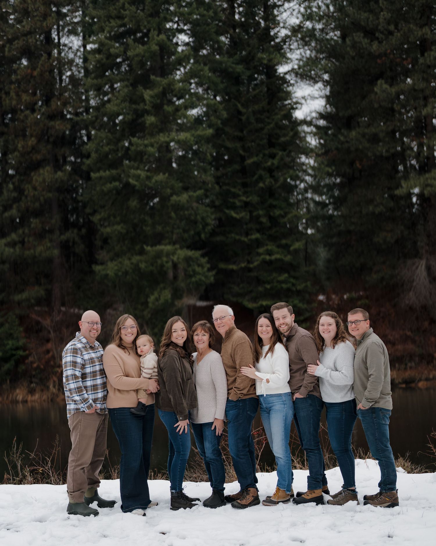 Family group portrait in snow with evergreen trees and pond backdrop, wearing casual winter attire.