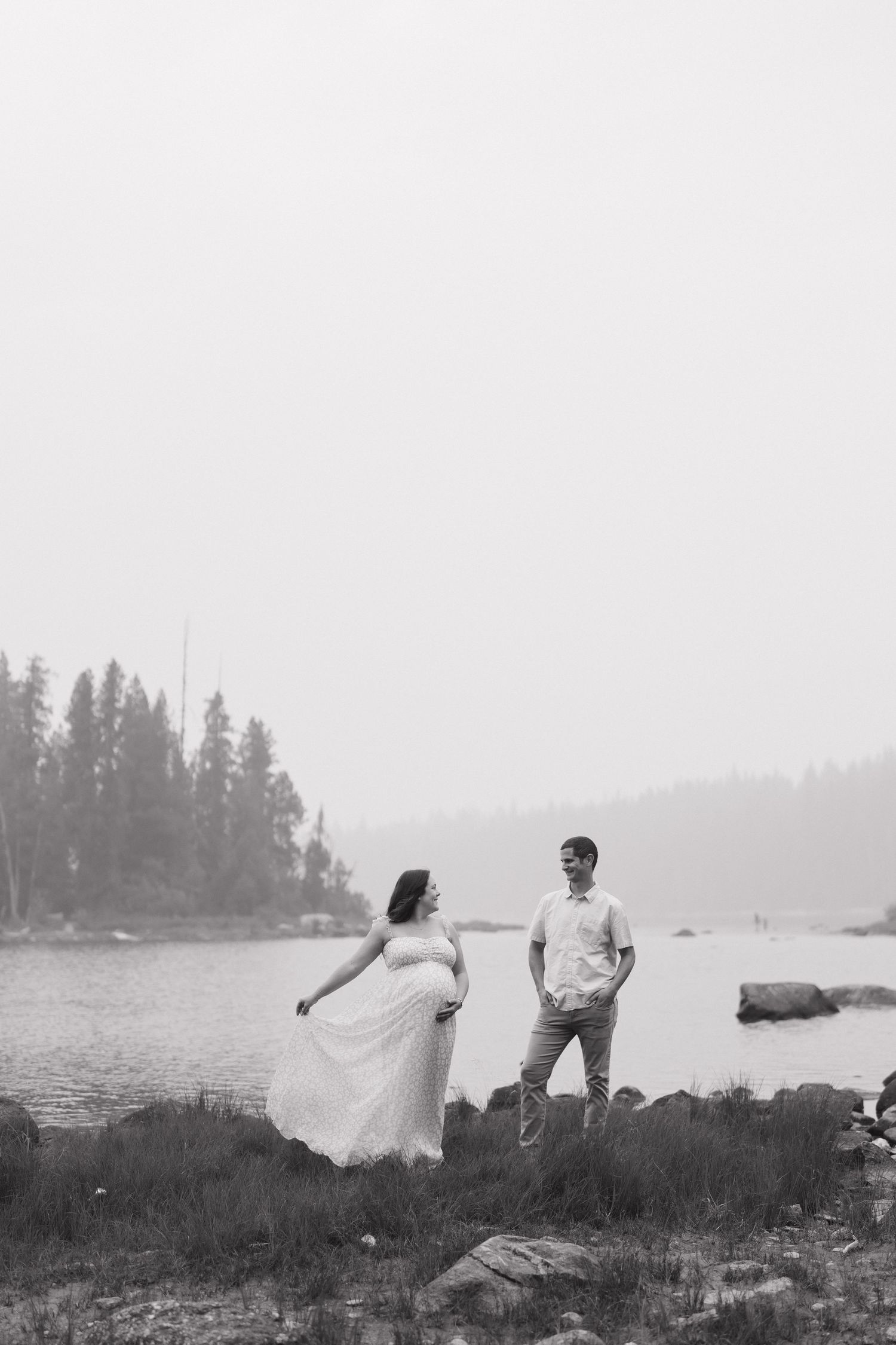 Black and white photo of a pregnant couple standing by a misty lake surrounded by pine trees.