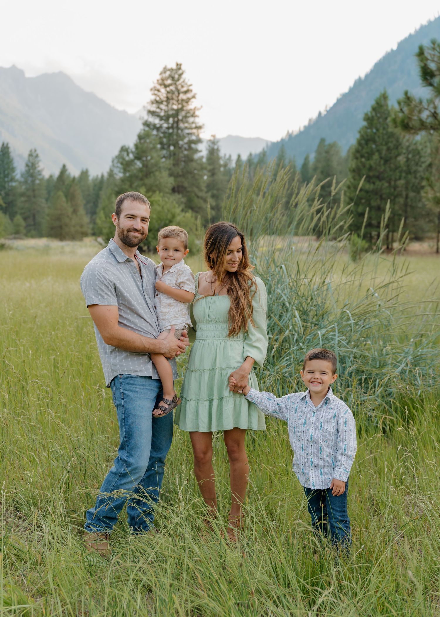Family of four posing in a lush green meadow with mountain and pine tree backdrop during golden hour.