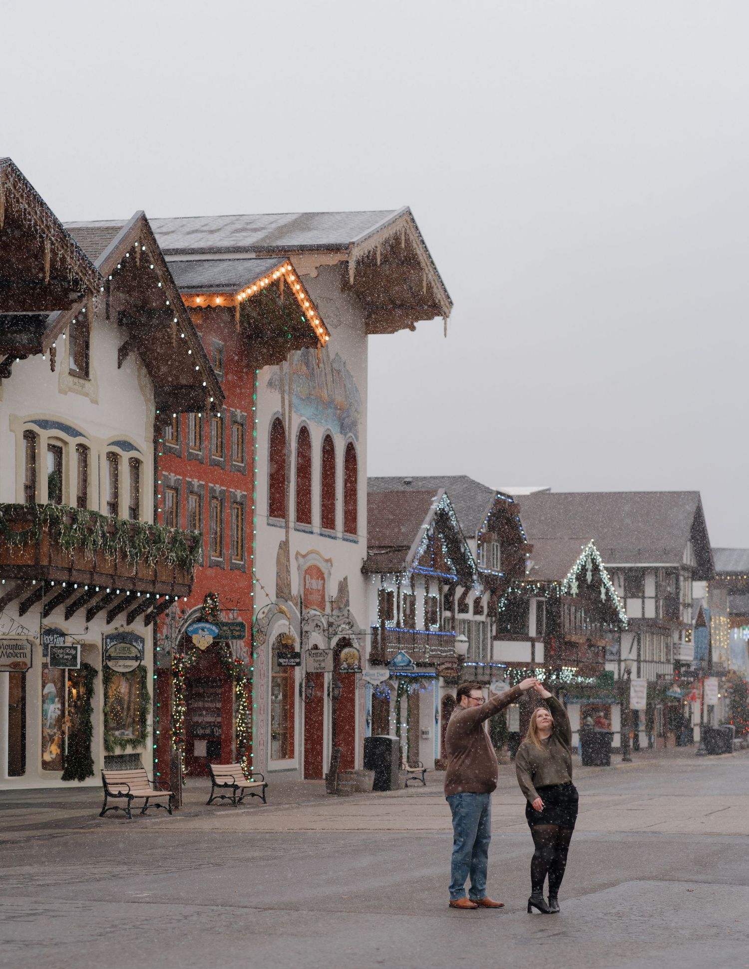 Couple strolling through a charming Bavarian-style village street decorated with Christmas lights on a snowy day.