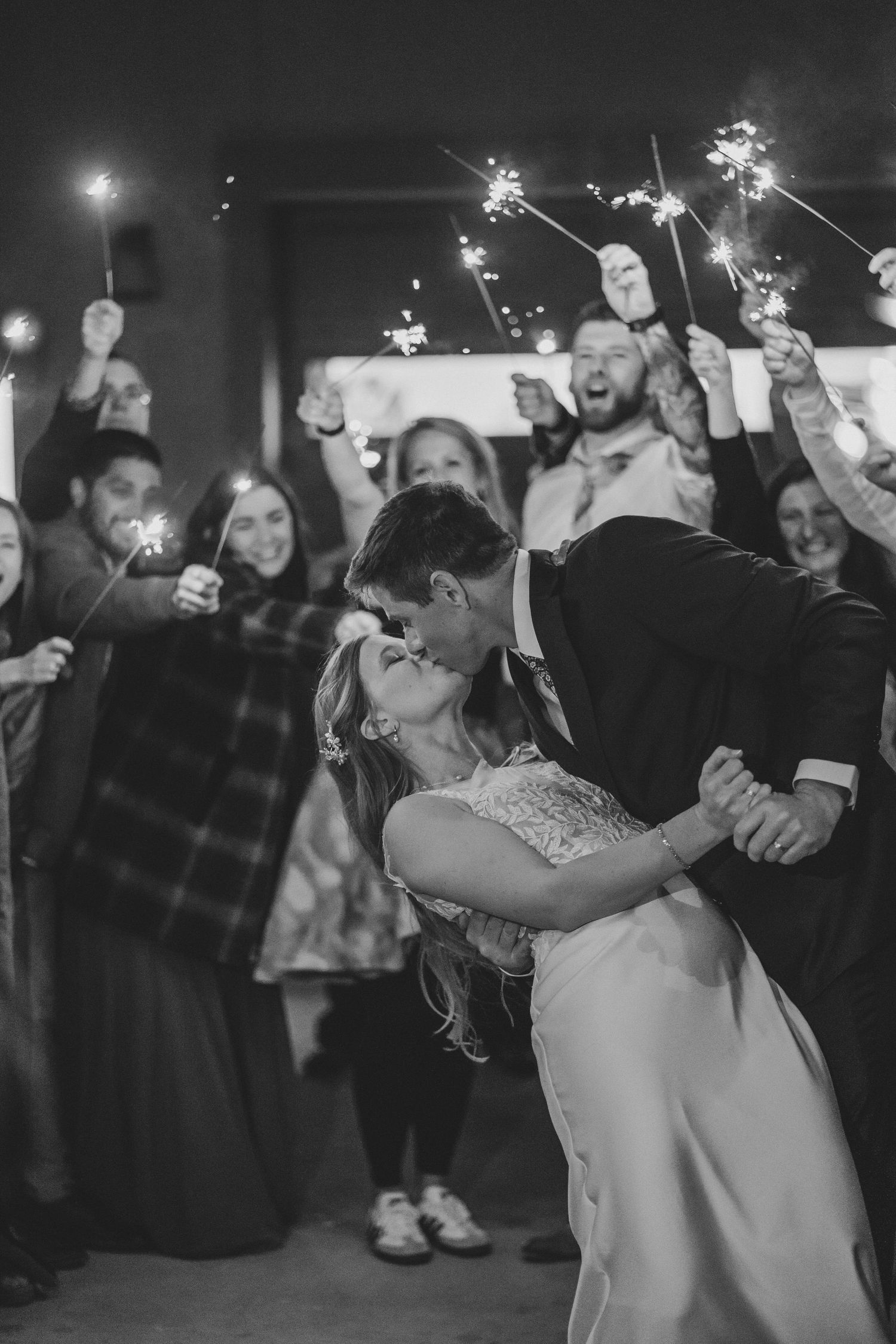 Couple shares romantic dip kiss while celebrating with sparklers at wedding reception in black and white.