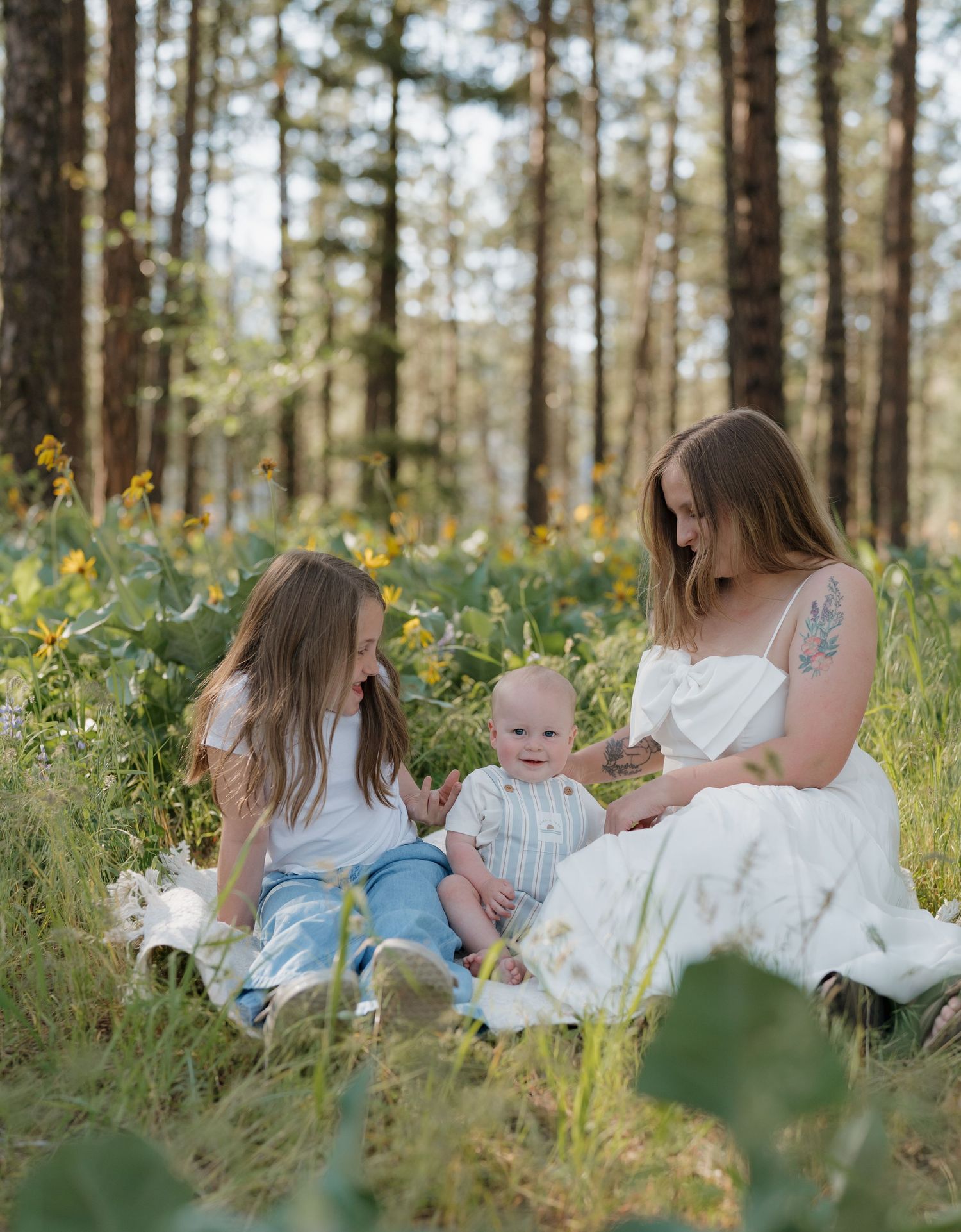A mother in a white dress sits with her two young children in a sunlit forest meadow with wildflowers.