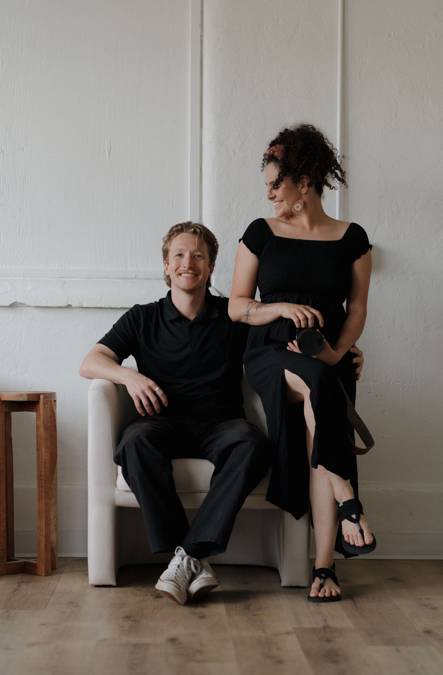 Two people in black clothing sit and interact playfully on a wooden floor against a white wall in a minimalist setting.