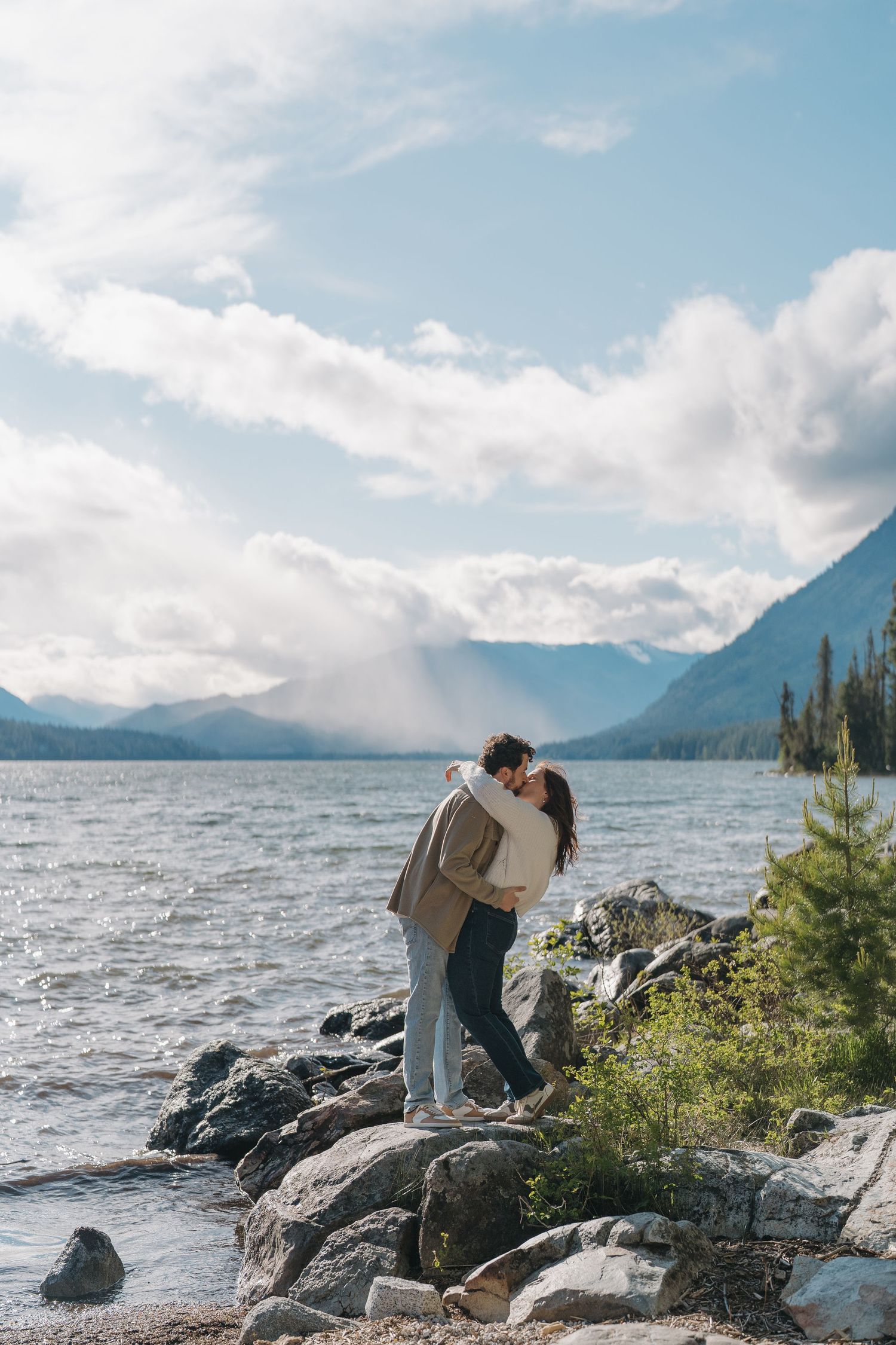 Couple kissing on rocky lakeshore with mountain backdrop and cloudy blue sky in a romantic outdoor setting.