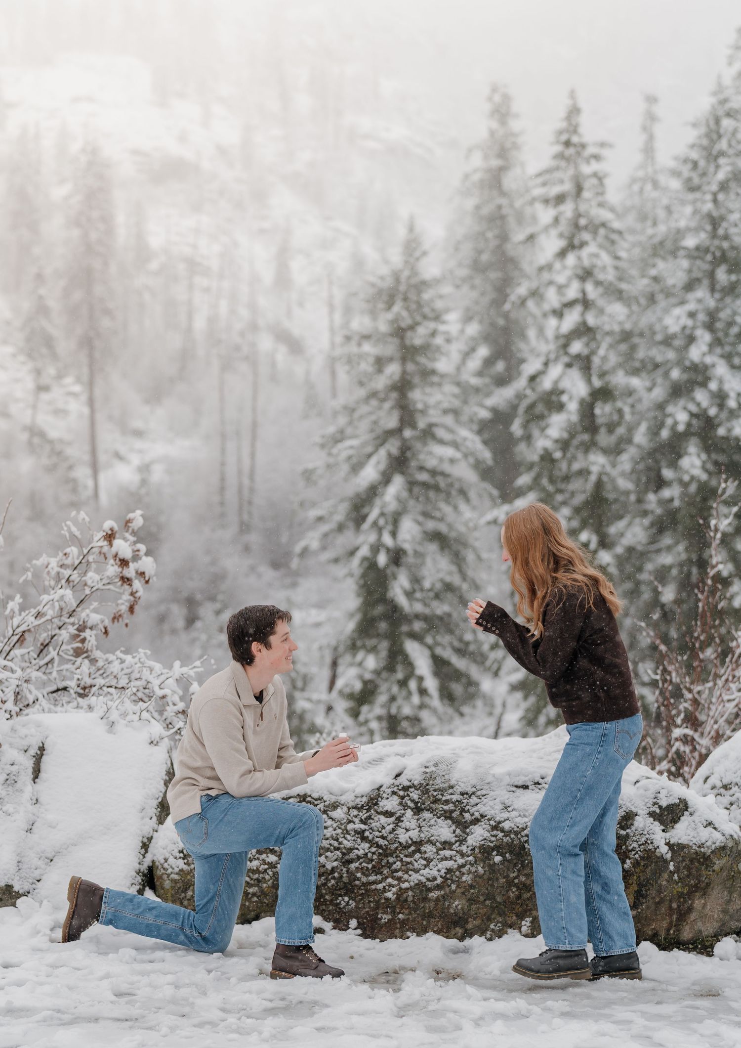 Man proposing on one knee to woman in snowy winter forest landscape with snow-covered pine trees.