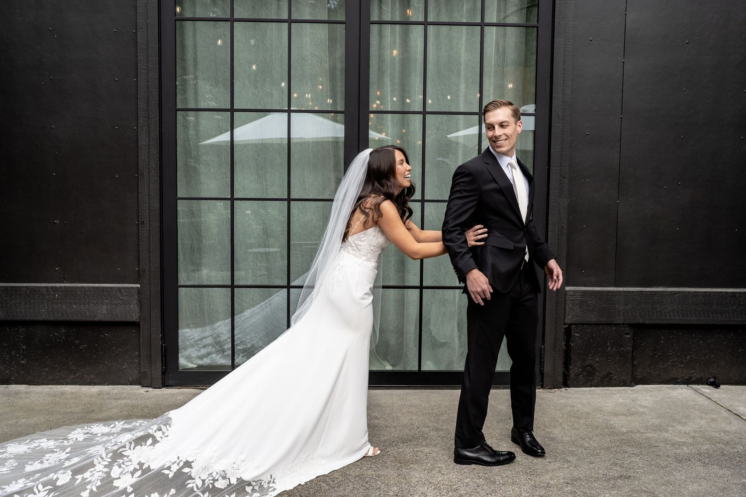 A bride in a white gown shares a playful moment with a groom before a glass door backdrop.