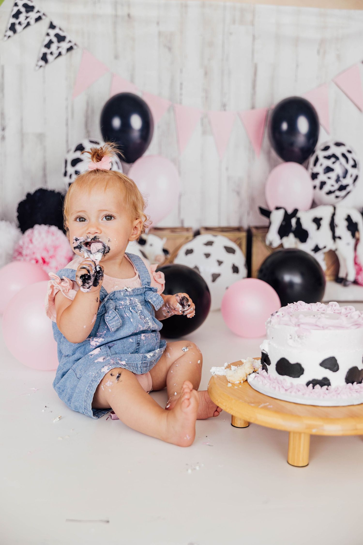 First birthday cake smash portrait of a baby surrounded by pink, black, and white balloons with a decorated birthday cake.