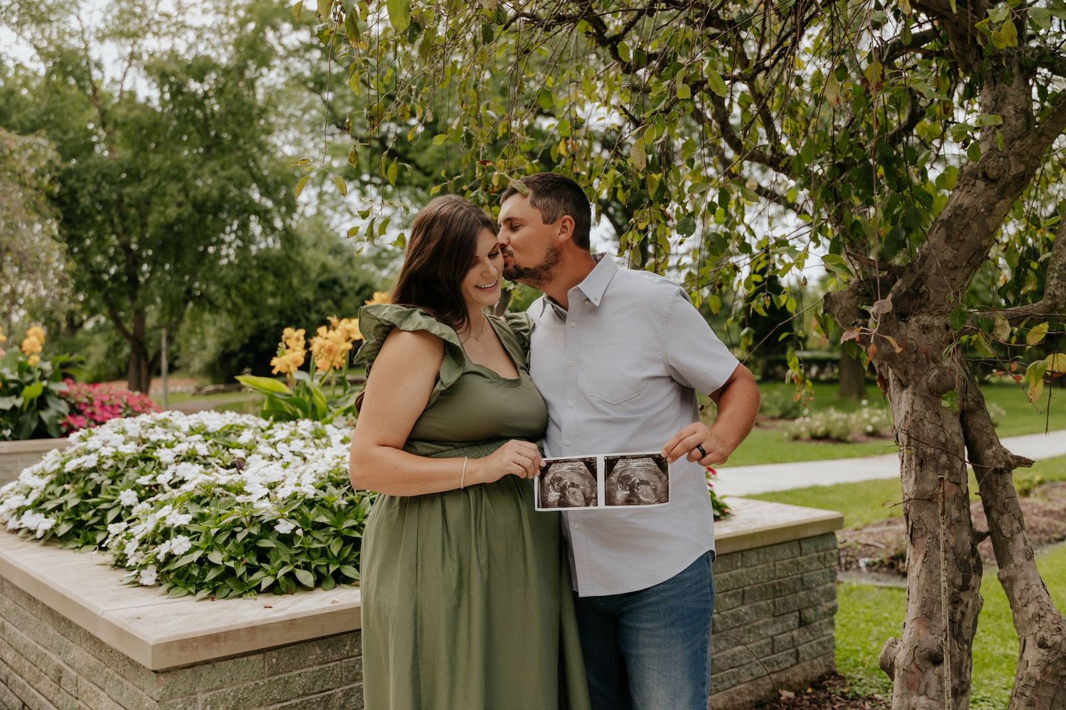 Outdoor maternity photoshoot of an expecting couple sharing a kiss beside a blooming flower garden, with the mother wearing a flowing green