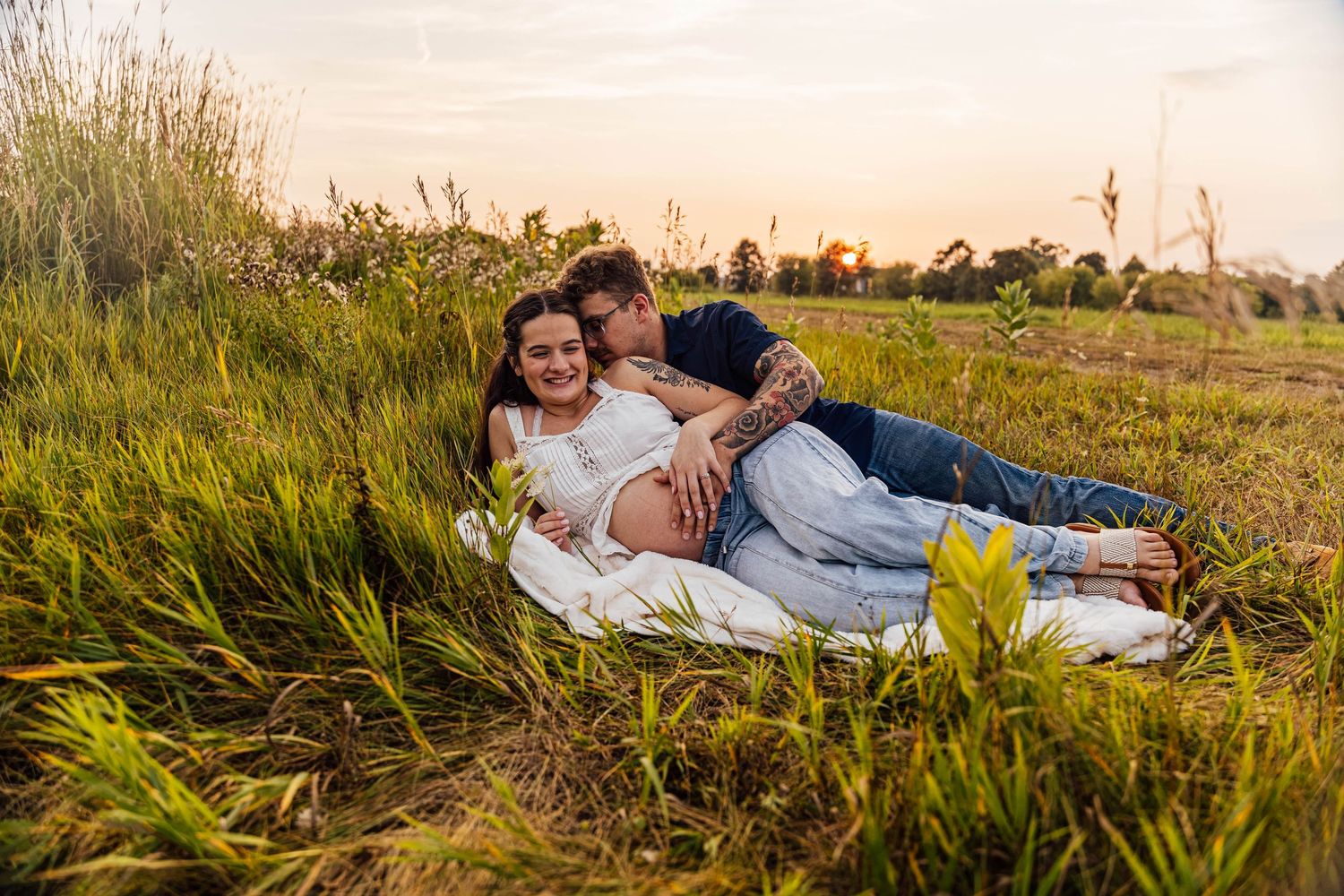 Outdoor maternity photoshoot of an expecting couple cuddling together in a grassy field during golden hour, with warm sunset light.