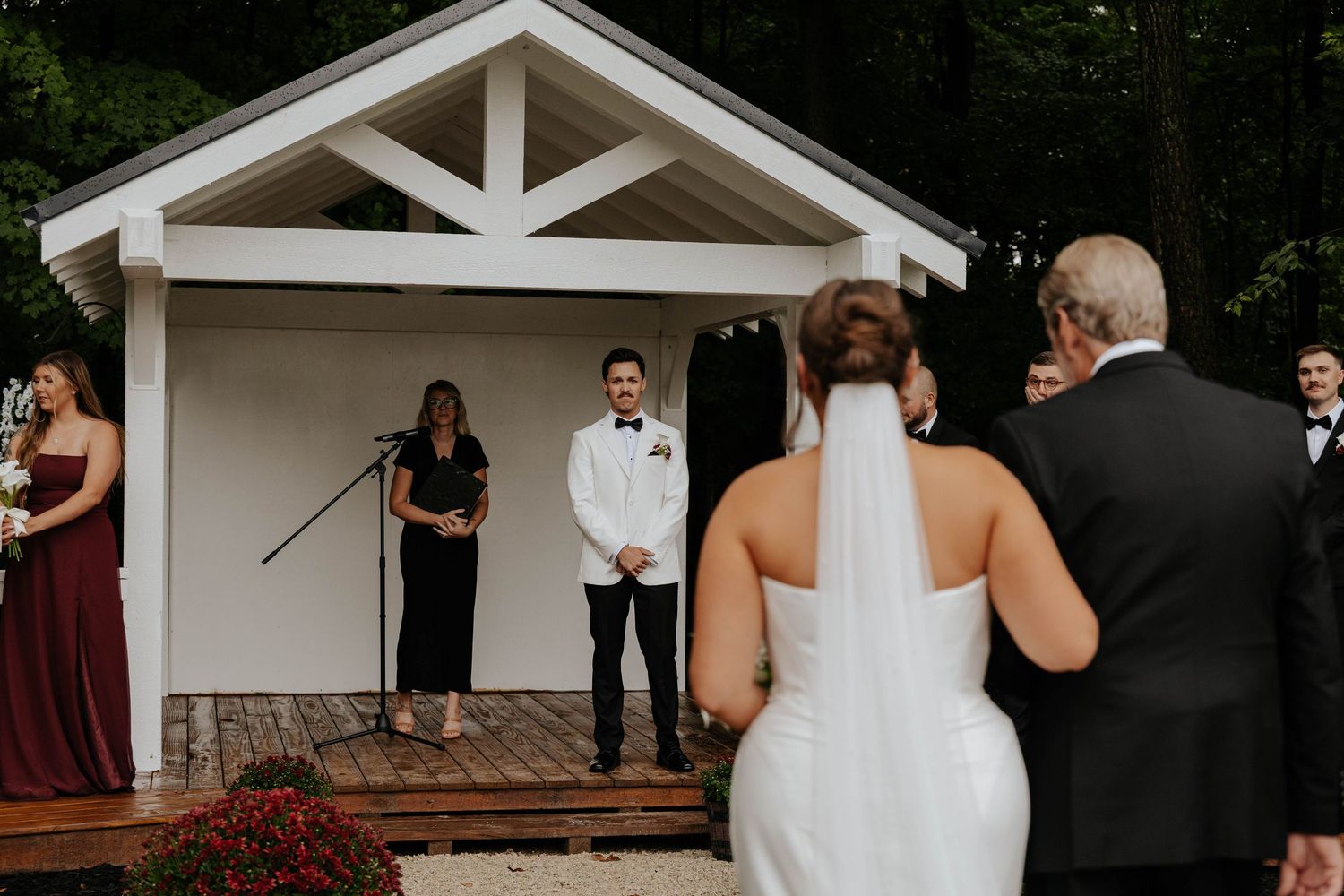 Bride walking down the aisle with her escort toward an outdoor wedding ceremony beneath a white wooden arbor in Battle Creek, Michigan.