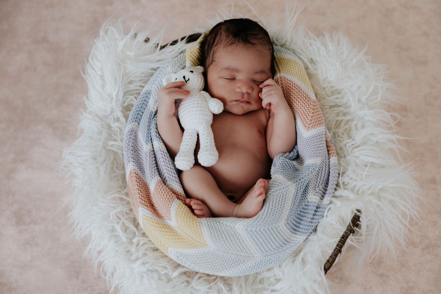 Overhead newborn portrait of a sleeping baby curled up in a soft cream wrap, cuddling a small stuffed bunny in a fluffy basket.