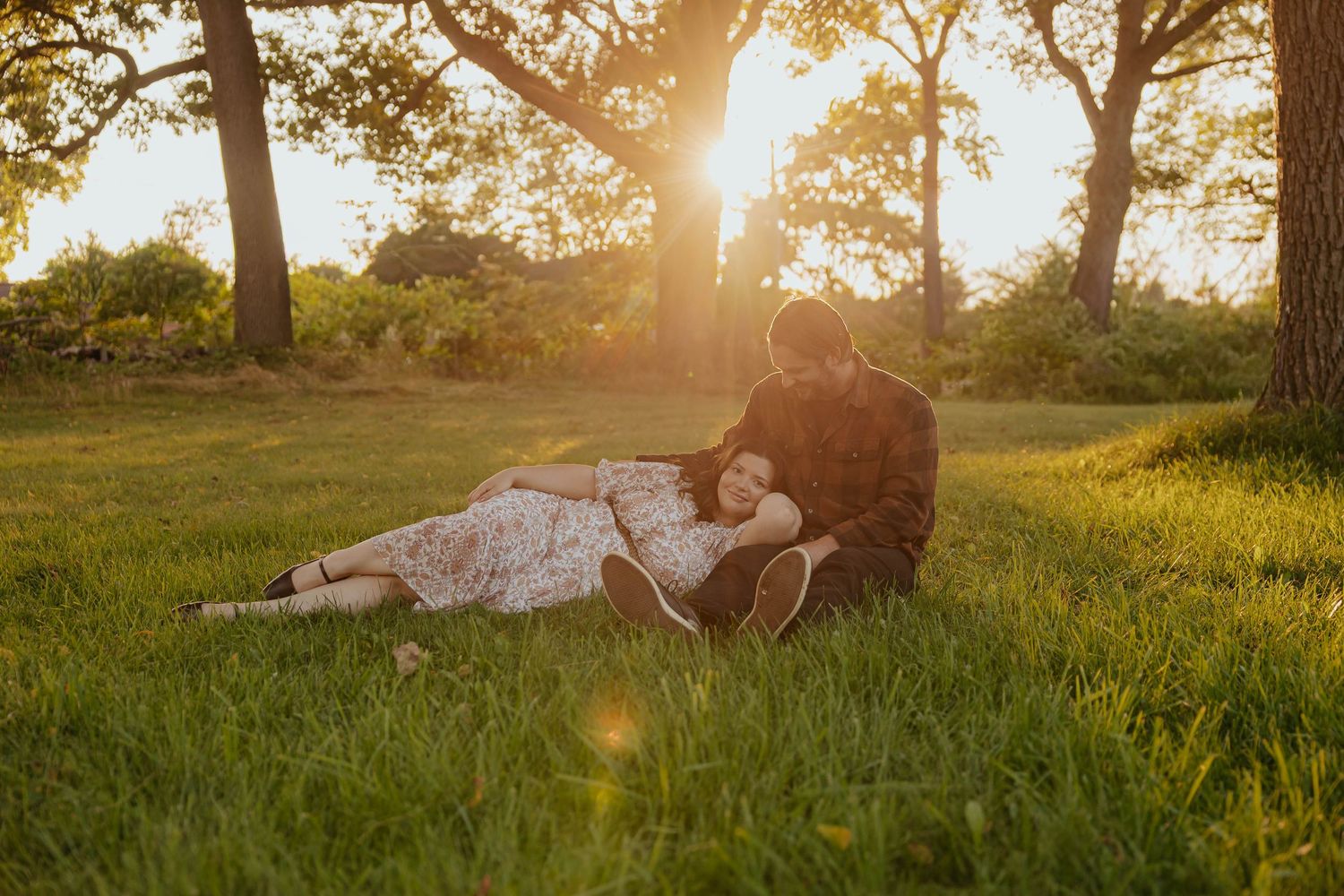 Golden hour engagement photos of a couple cuddling in a grassy field beneath the trees near Lake Odessa, Michigan.