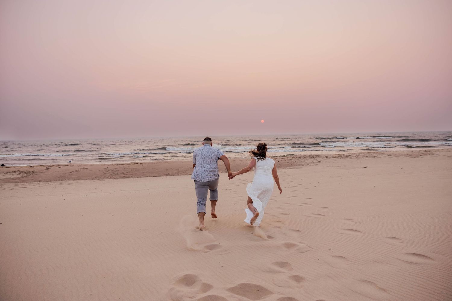 Couple running hand in hand toward Lake Michigan during a sunset beach engagement session in Saugatuck, Michigan.