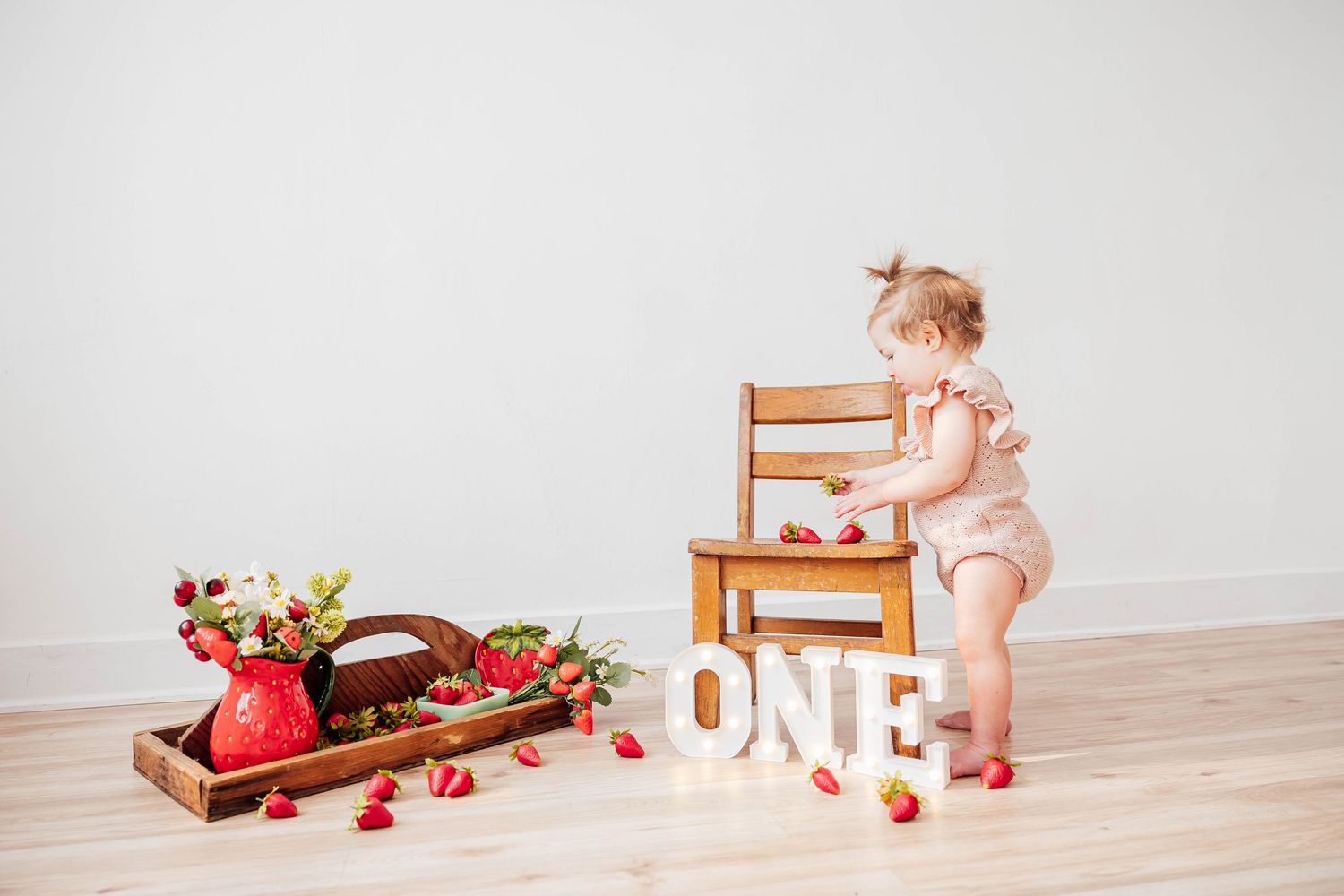 First birthday cake smash photoshoot featuring a baby girl standing beside a wooden high chair, strawberries, and a ONE birthday sign.