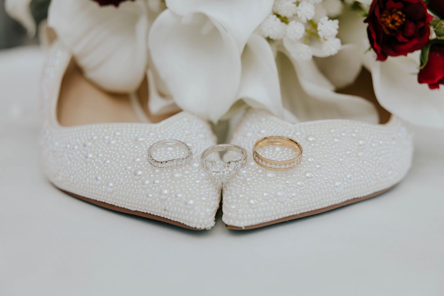 Elegant wedding detail photo of gold wedding rings resting on sparkling bridal shoes beside white flowers in Battle Creek, Michigan.