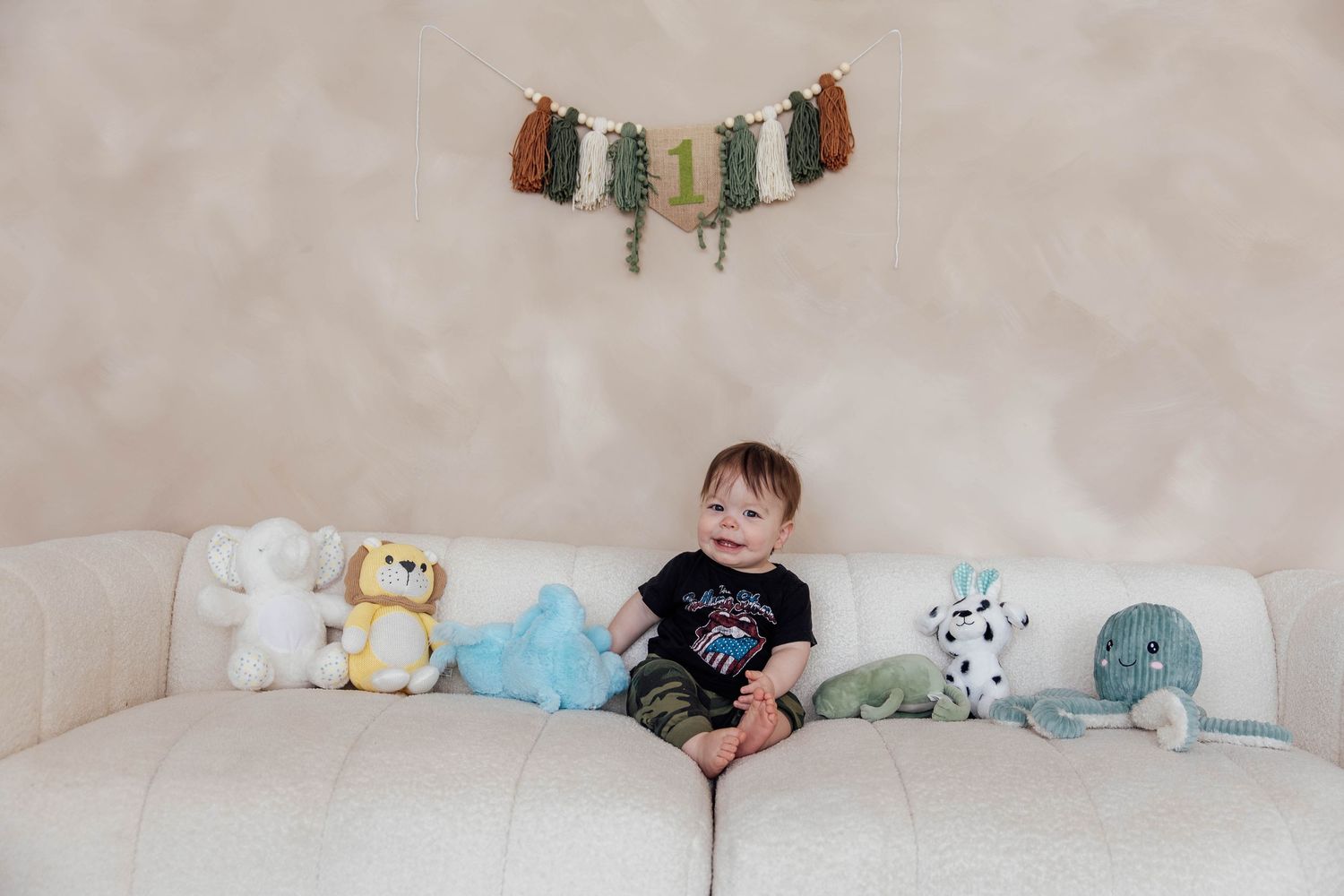 First birthday milestone portrait of a baby boy sitting on a cream-colored couch surrounded by soft blue decorations and stuffed animals.