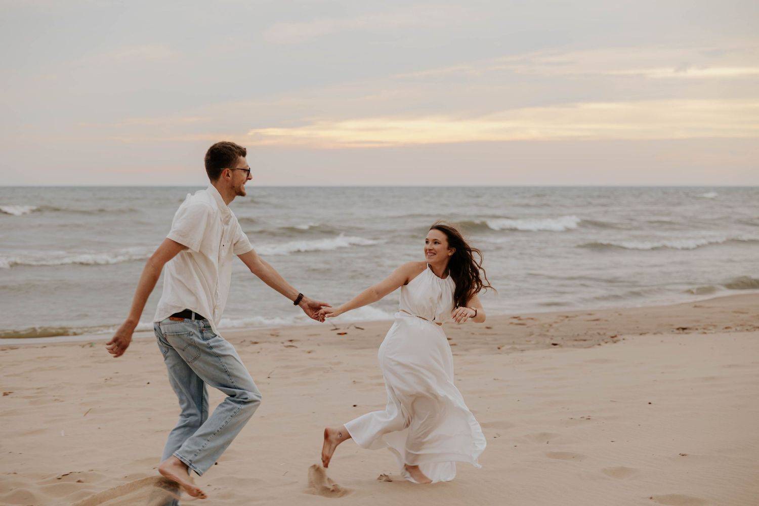 Playful beach engagement photos of a couple running hand in hand along the Lake Michigan shoreline in Holland, Michigan.