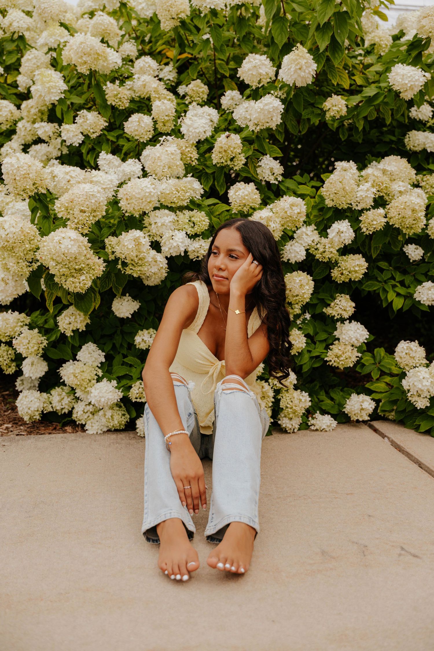 Outdoor senior portrait of a young woman sitting in front of blooming white hydrangeas, wearing a casual summer outfit.