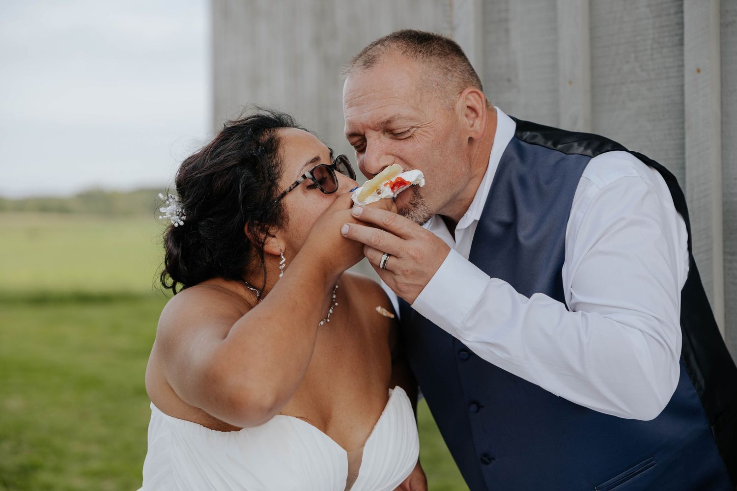 Candid wedding reception photo of a bride and groom sharing a playful bite of cake beside a rustic barn in Sparta, Michigan.