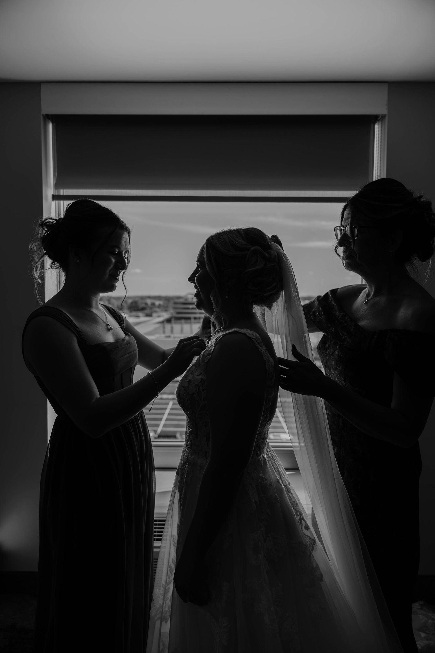 Black and white silhouette of bride being helped into wedding dress by two bridesmaids in front of a bright window in Lansing, Michigan.