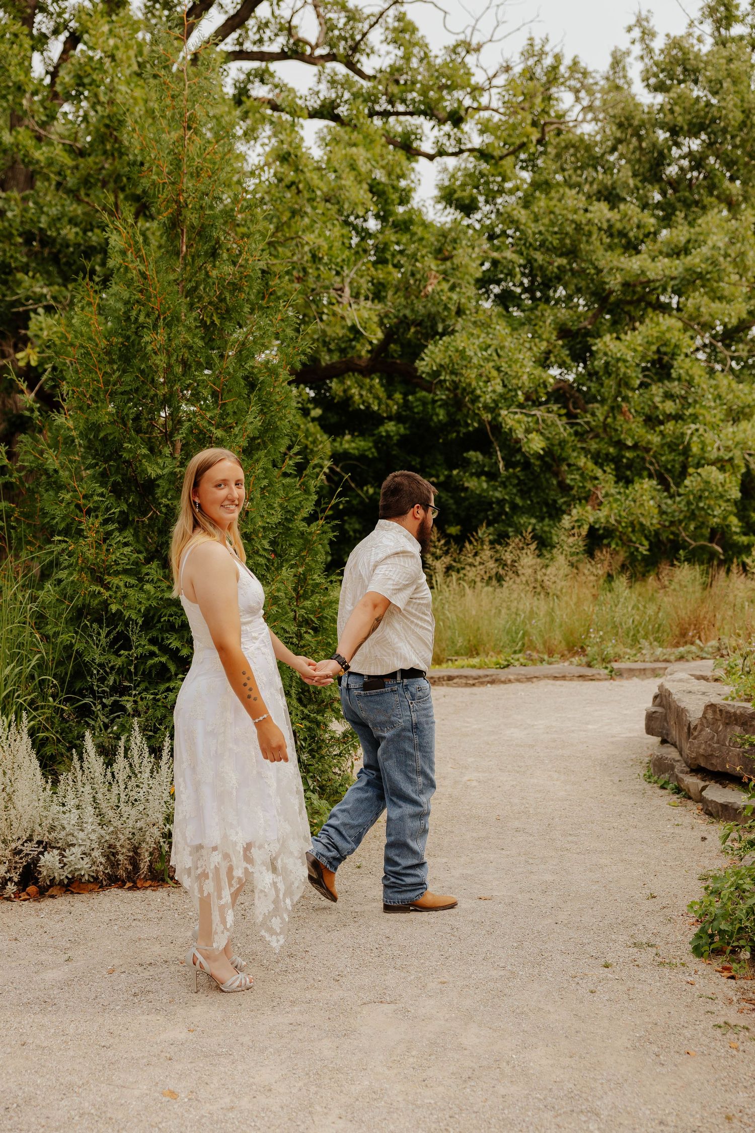 Natural engagement photography of a couple holding hands on a garden pathway surrounded by lush greenery in Michigan.