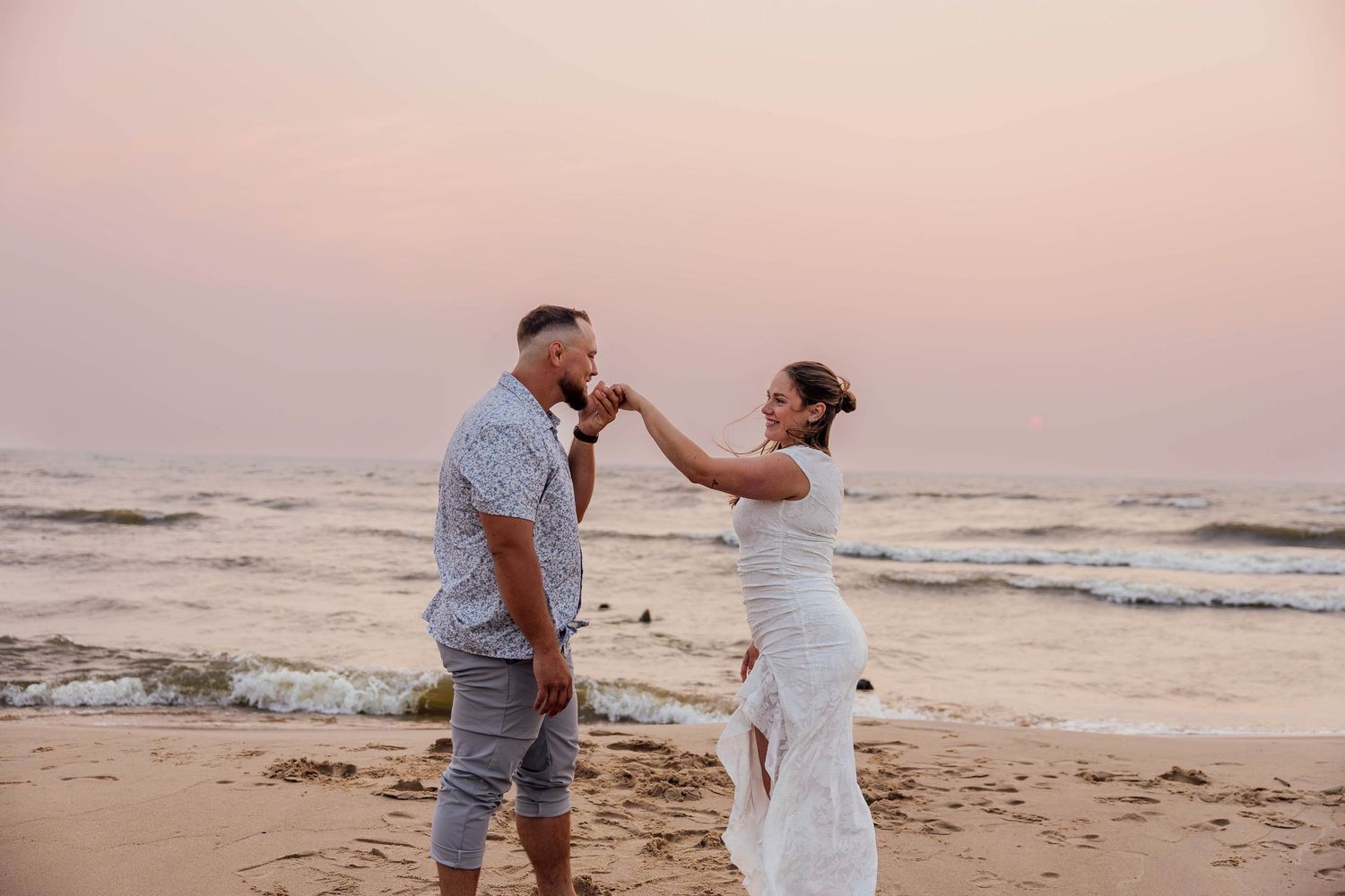 Romantic Saugatuck beach engagement photos of a couple holding hands by Lake Michigan at sunset, with soft pink skies and gentle waves.