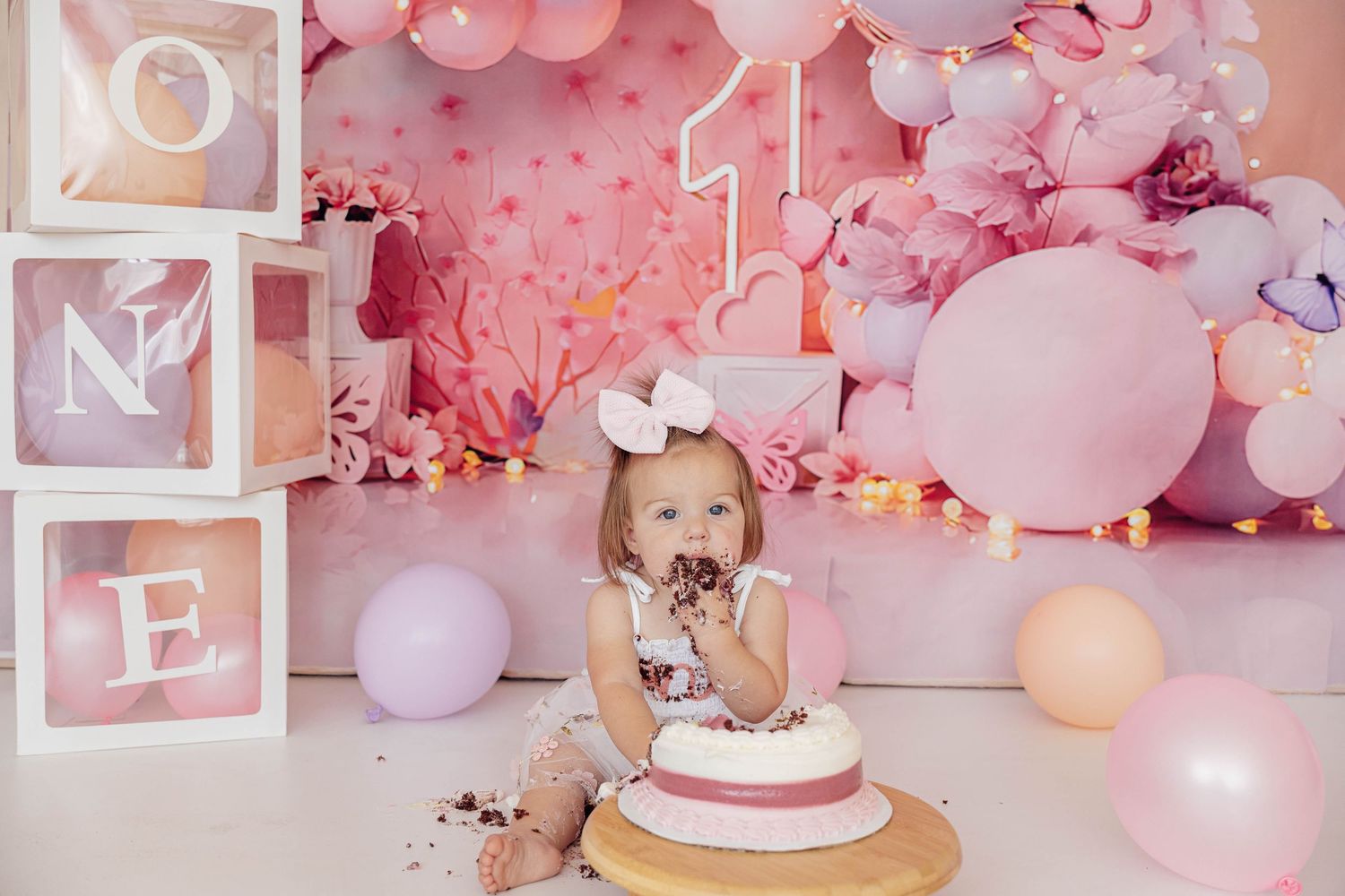 Pink first birthday cake smash photoshoot featuring a baby girl with a bow, birthday cake, pastel balloons, and floral decorations.