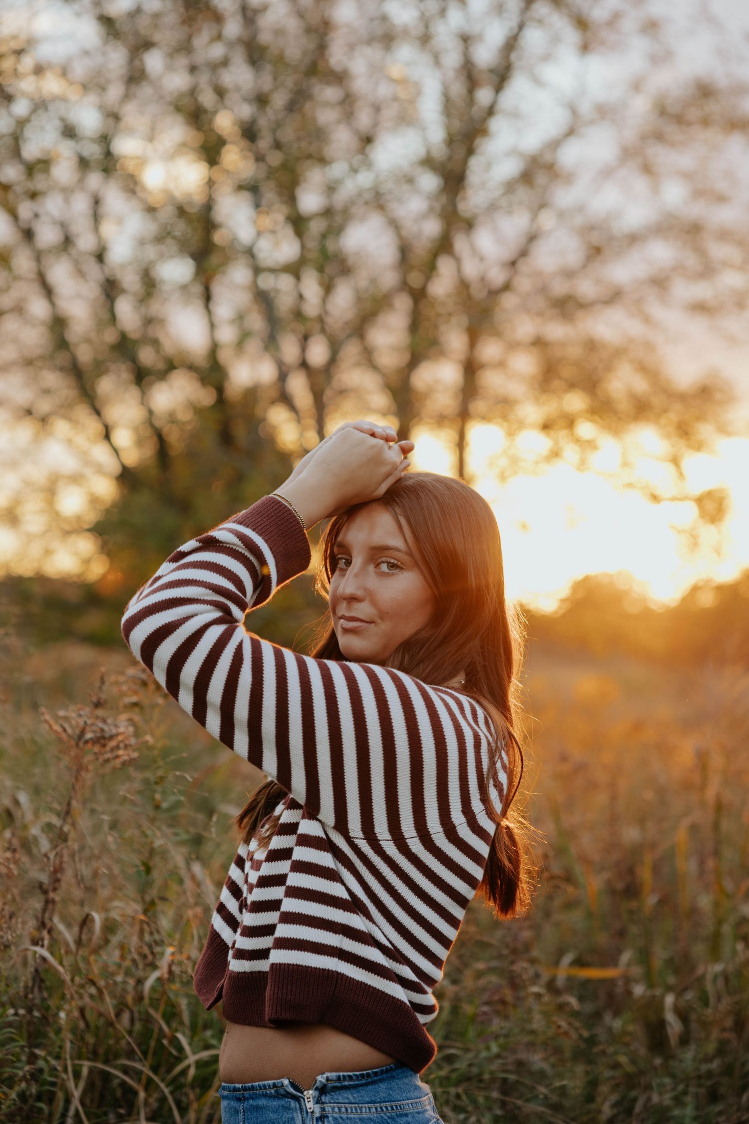 Golden-hour fall senior portrait of a young woman in a striped sweater posing in a sunlit field near Kalamazoo, Michigan.