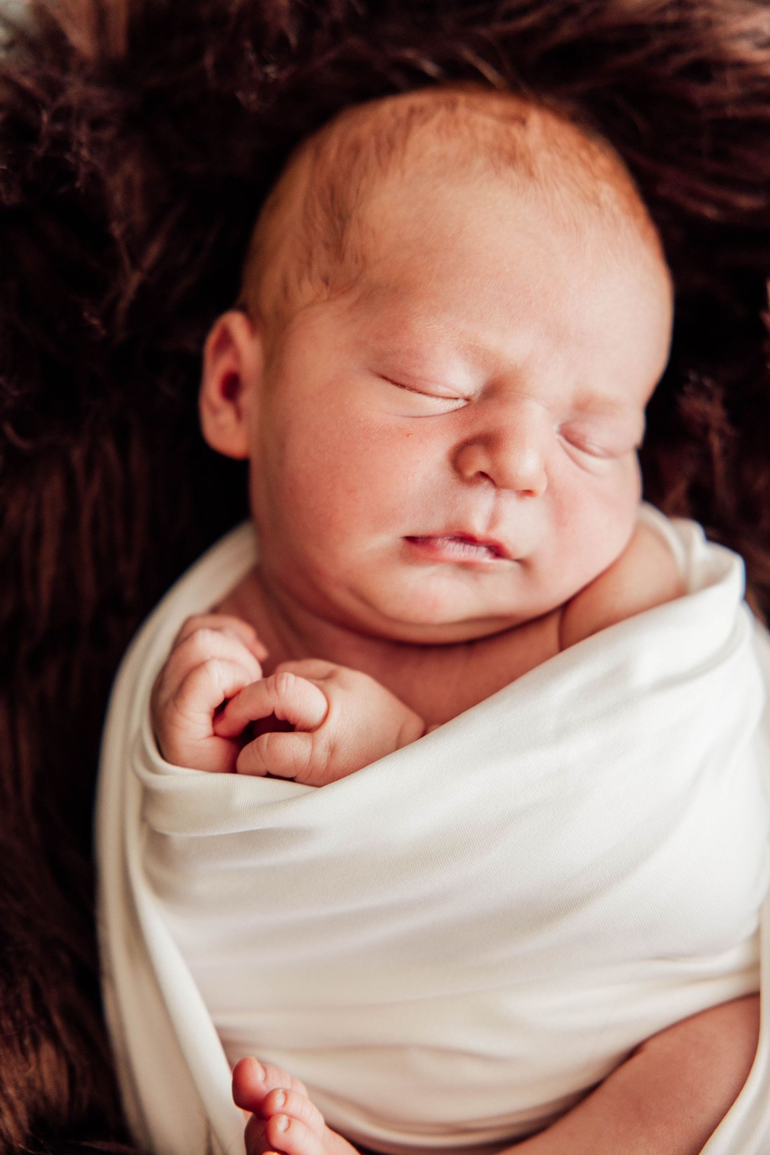 Close-up newborn portrait of a peacefully sleeping baby swaddled in a cream blanket against a soft, dark textured background.